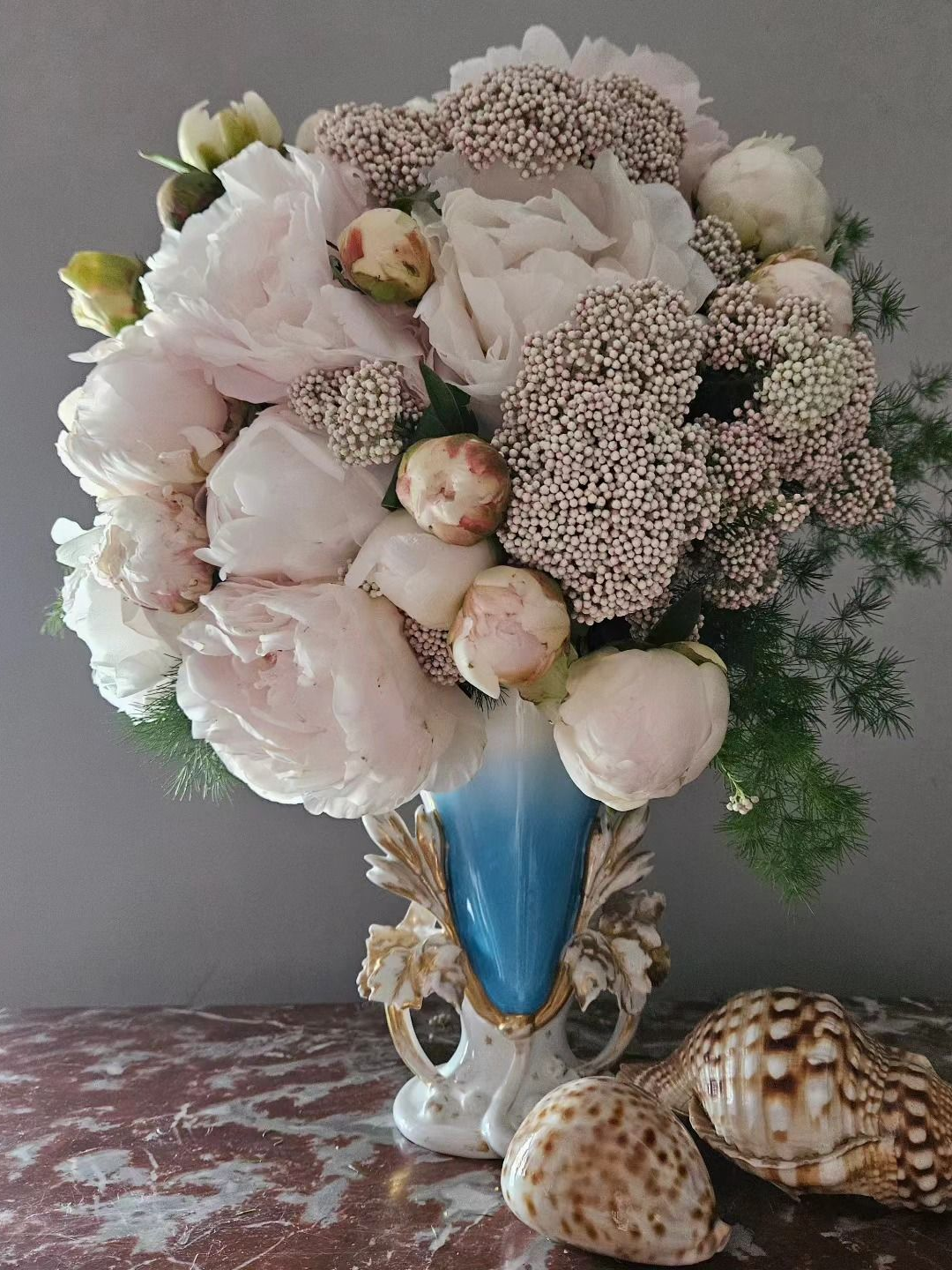 Bouquet de fleurs rose pâle et blanches dans un vase bleu, avec des coquillages sur une surface en marbre