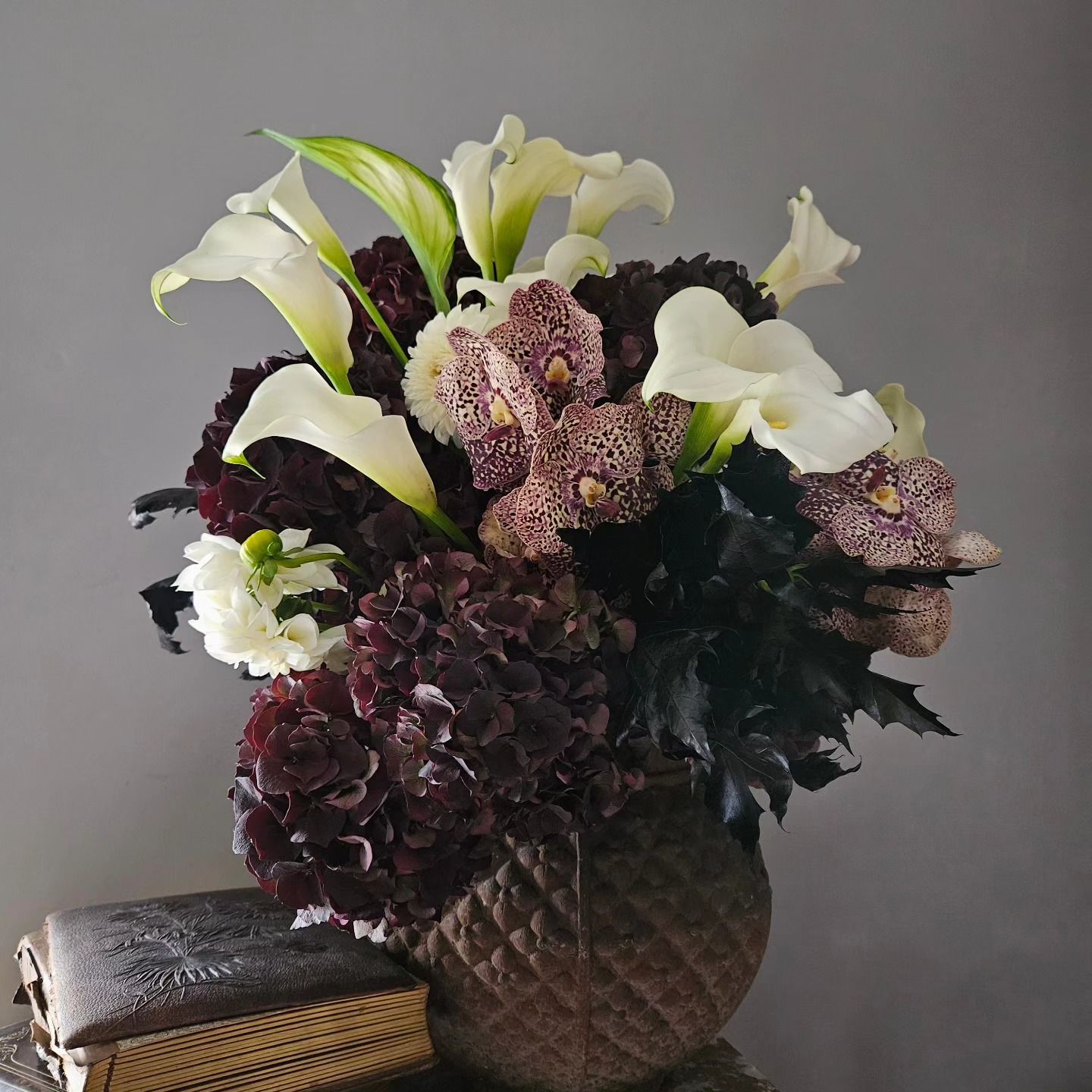 Bouquet de lys blancs et de fleurs violet foncé dans un vase texturé, à côté d'une pile de livres