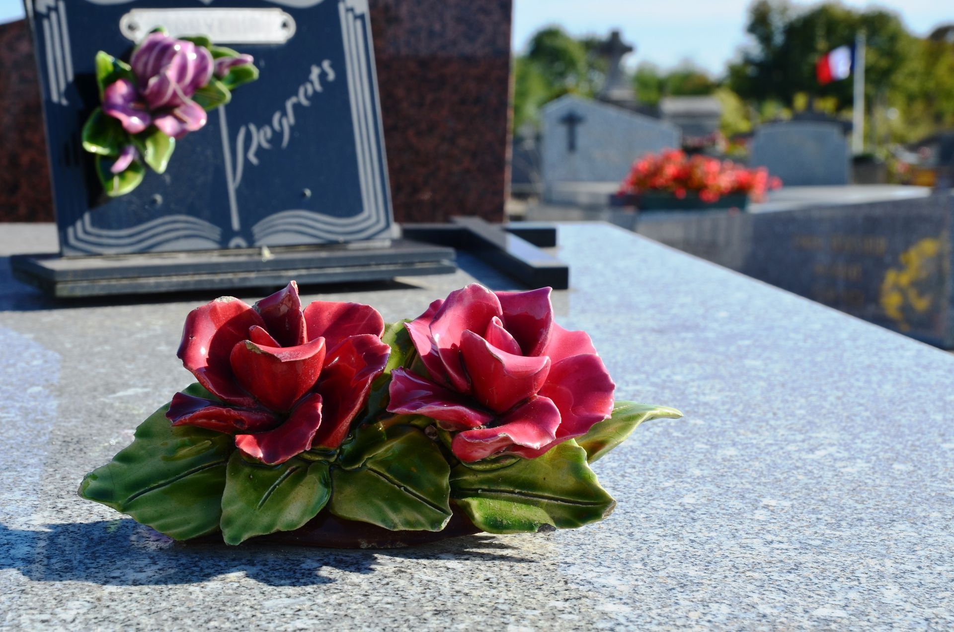 Des roses rouges sur une pierre tombale polie dans un cimetière, avec une plaque commémorative florale en arrière-plan.