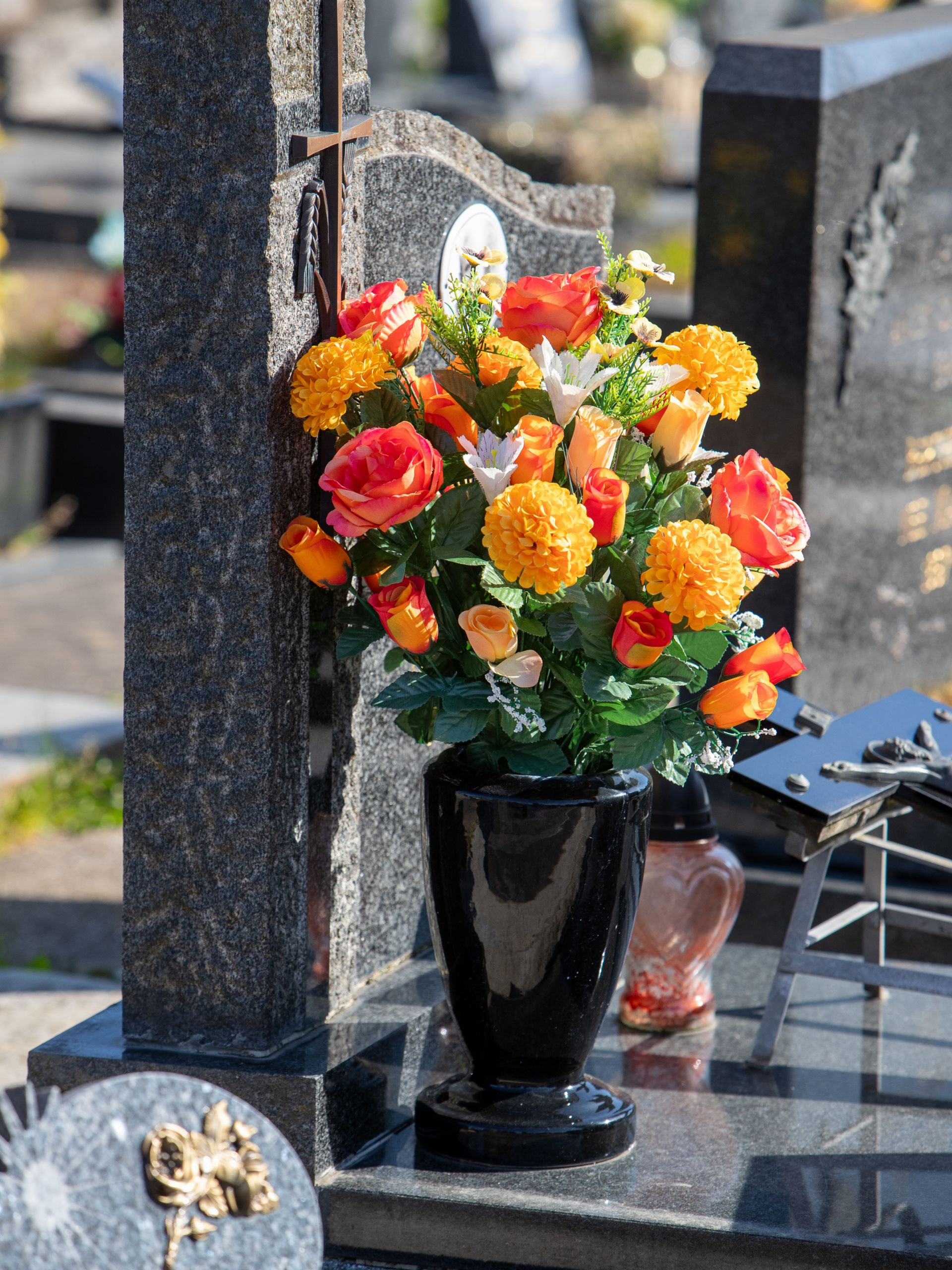 Des fleurs dans un vase noir, sur une tombe de cimetière, avec des fleurs orange et jaunes sur une pierre polie.