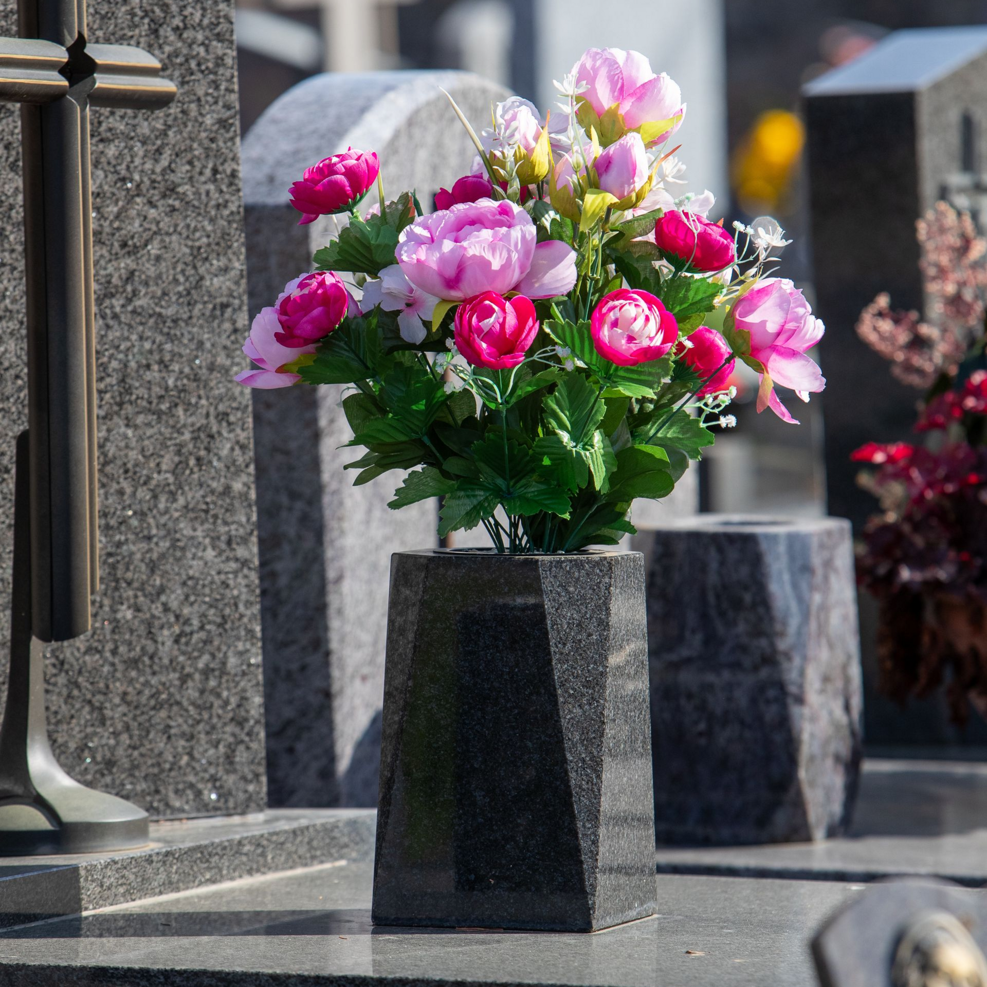 Des fleurs roses et blanches disposées dans un vase de granit au milieu des pierres tombales d'un cimetière.