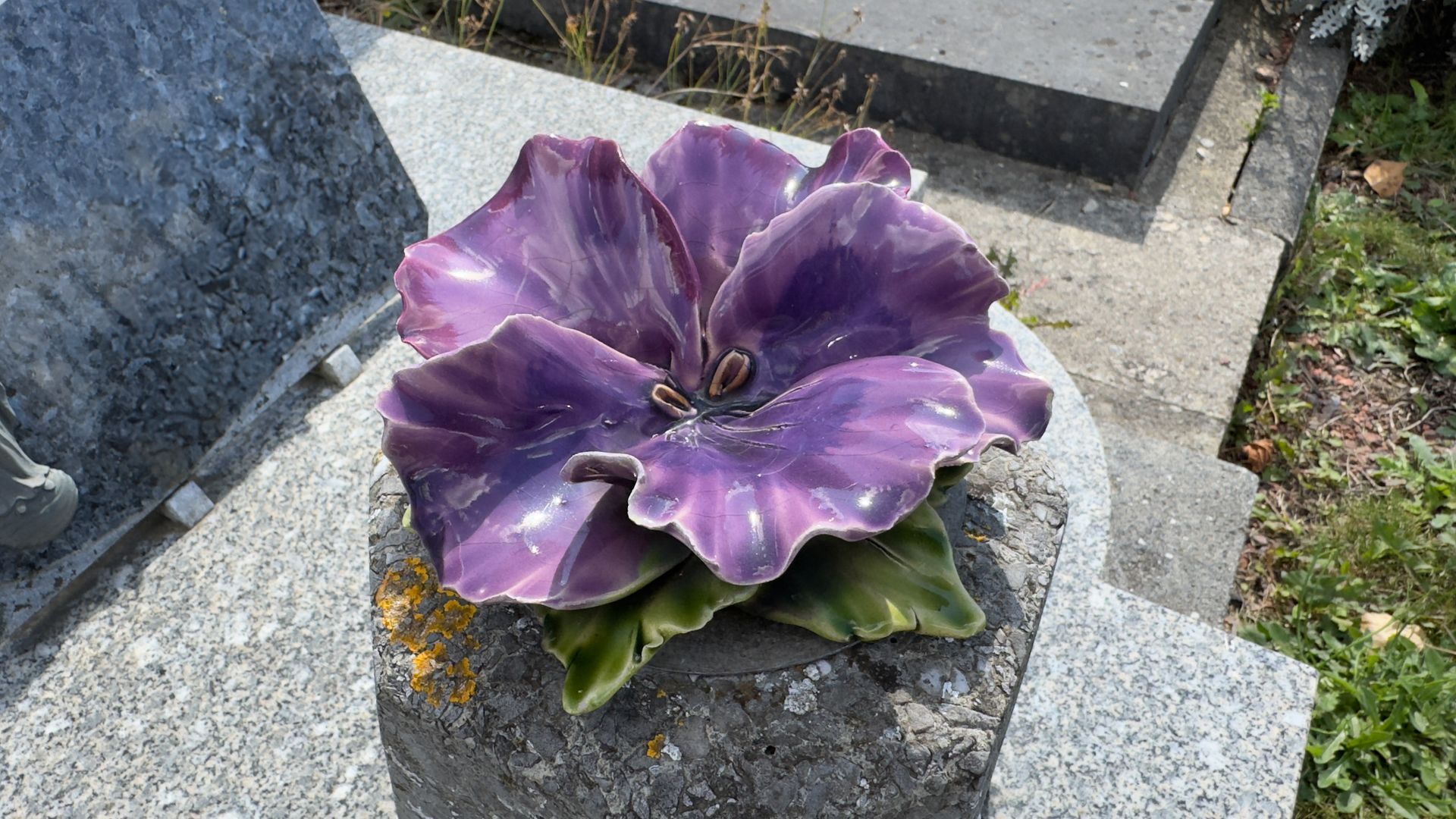 Fleur artificielle violette sur une stèle funéraire en granit gris dans un cimetière.