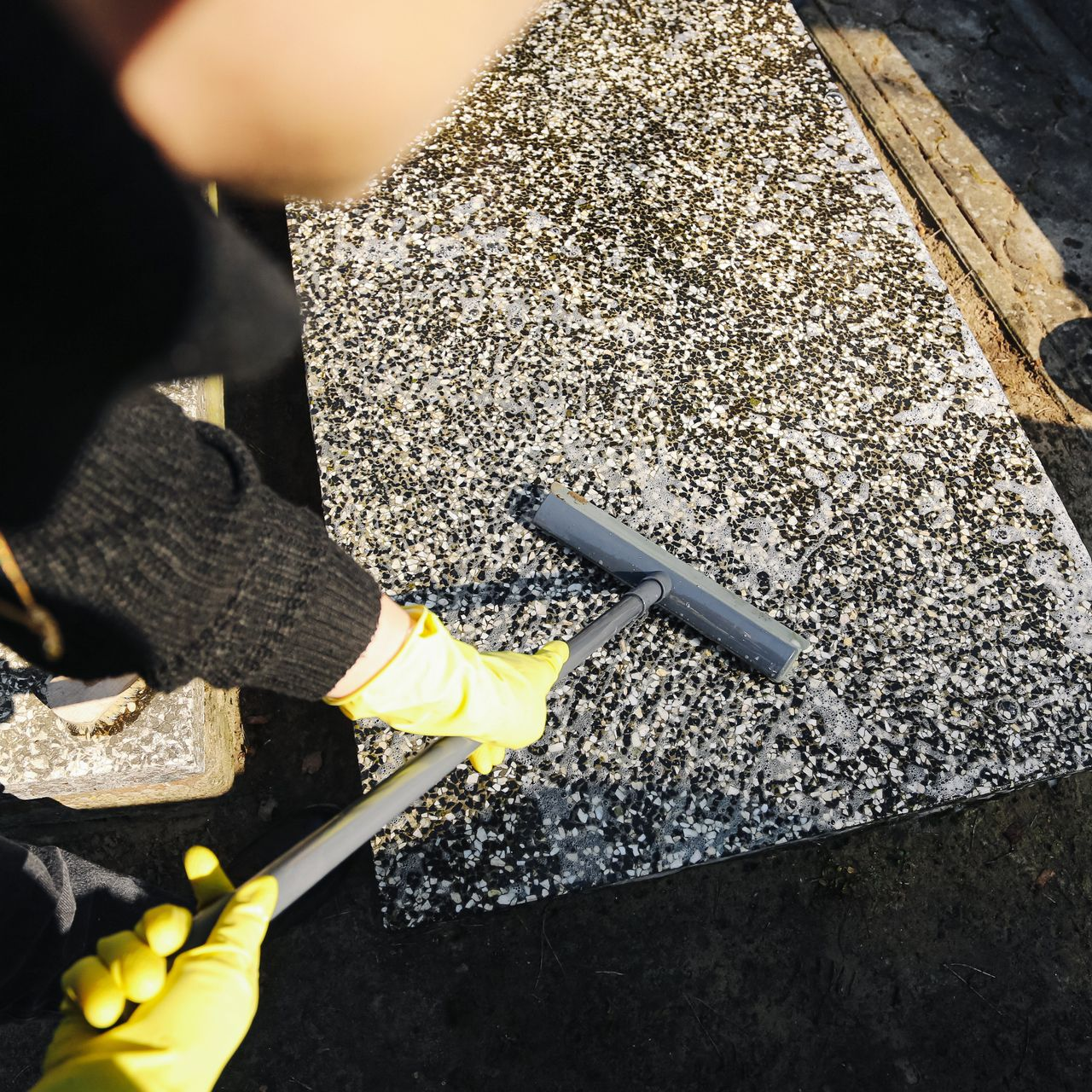 Une personne nettoie à haute pression une surface en béton mouchetée avec un tuyau noir et des gants jaunes.
