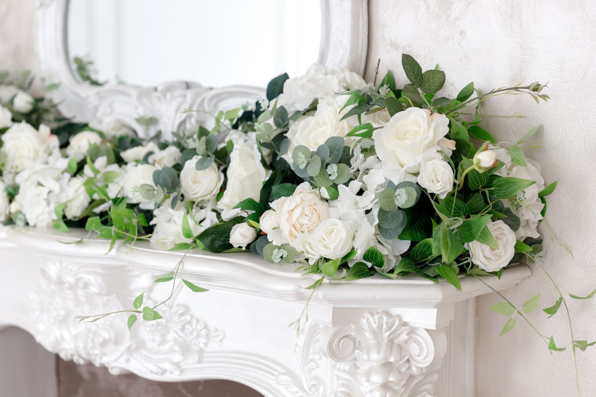 Des roses blanches et de la verdure disposées sur une cheminée blanche ornée, sous un miroir rond.