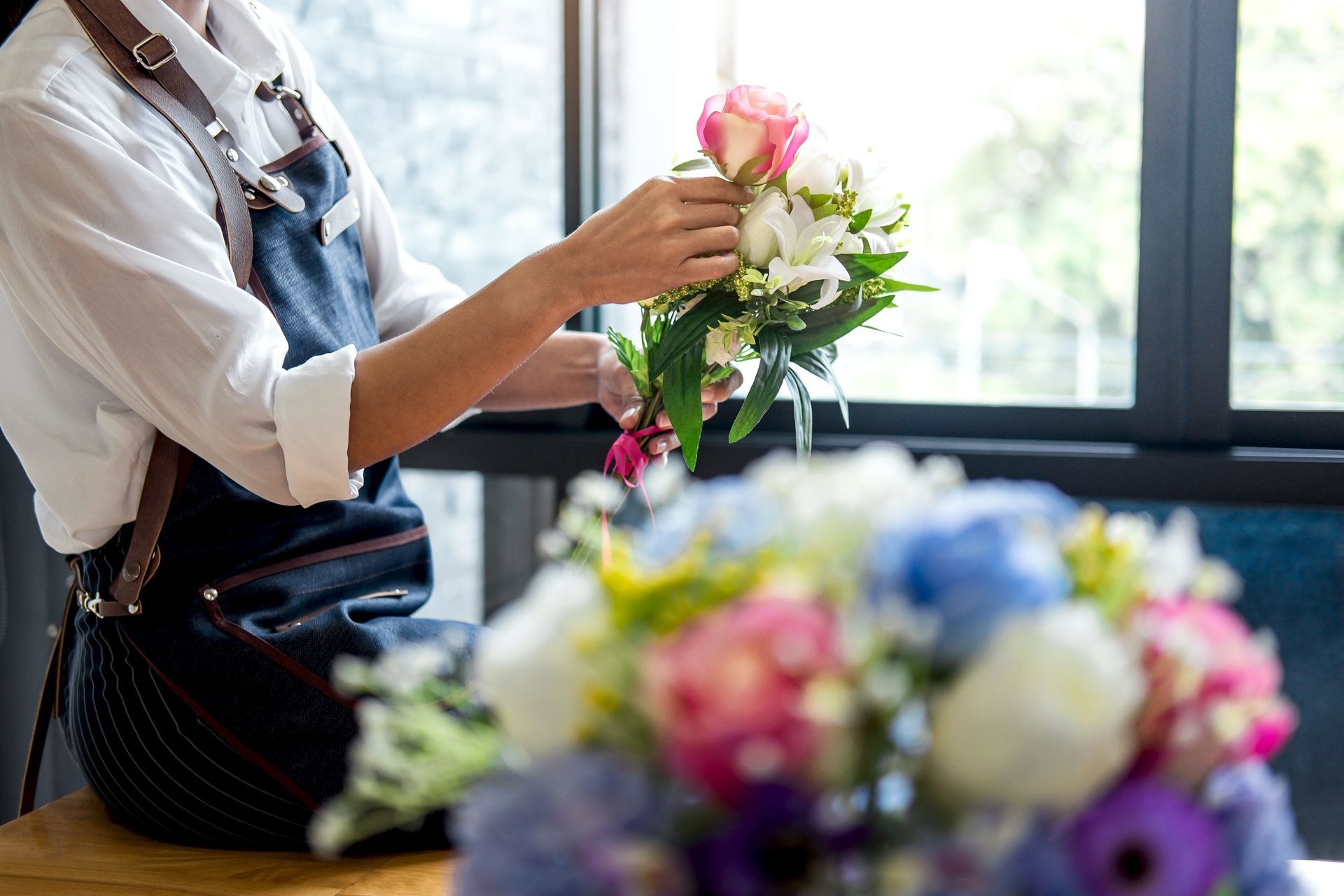 Une fleuriste arrange des fleurs roses et blanches sur une table de travail près d'une fenêtre lumineuse.