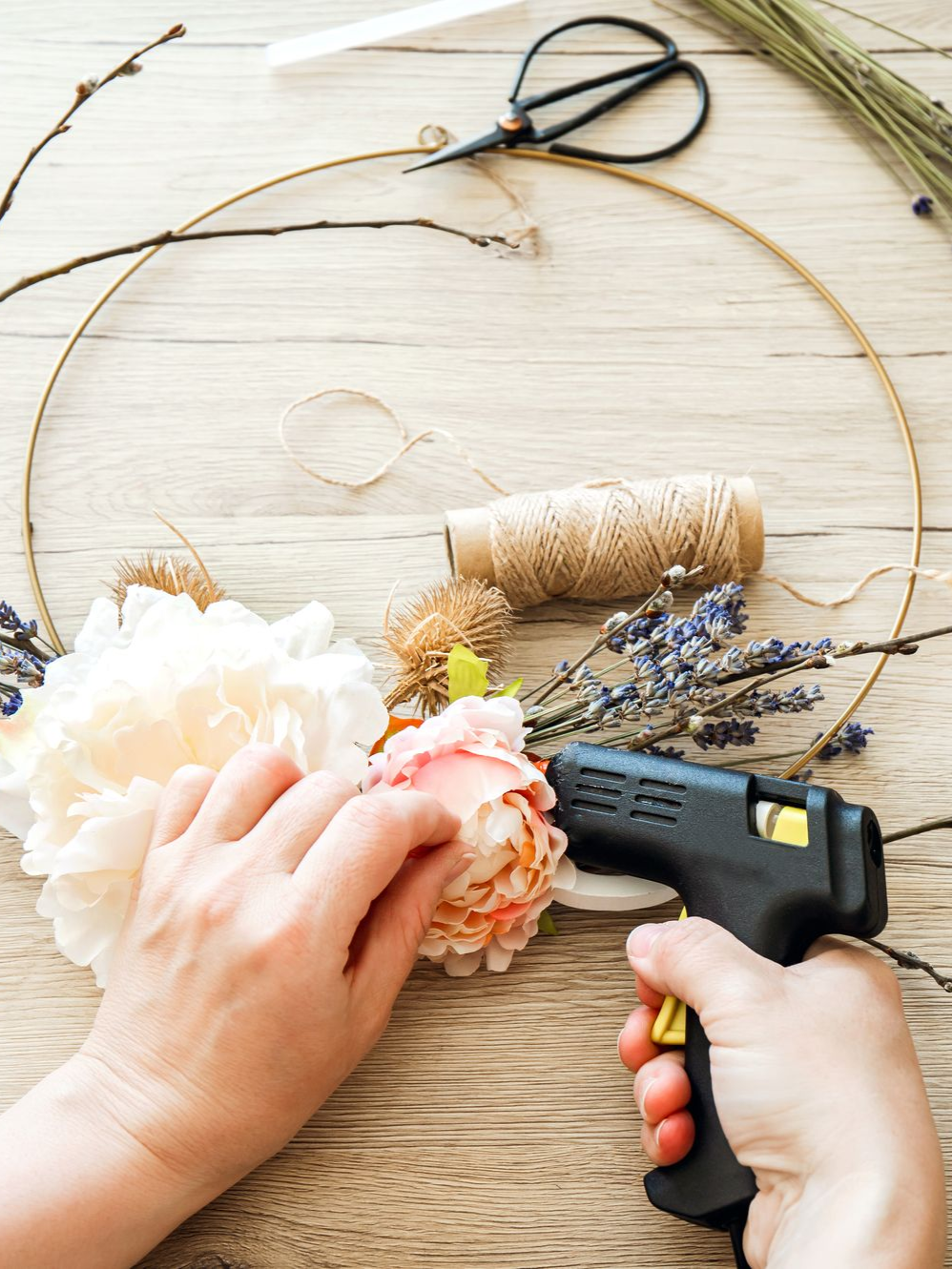 Des mains collent des fleurs et de la ficelle sur une couronne en forme de cercle posée sur une table en bois.