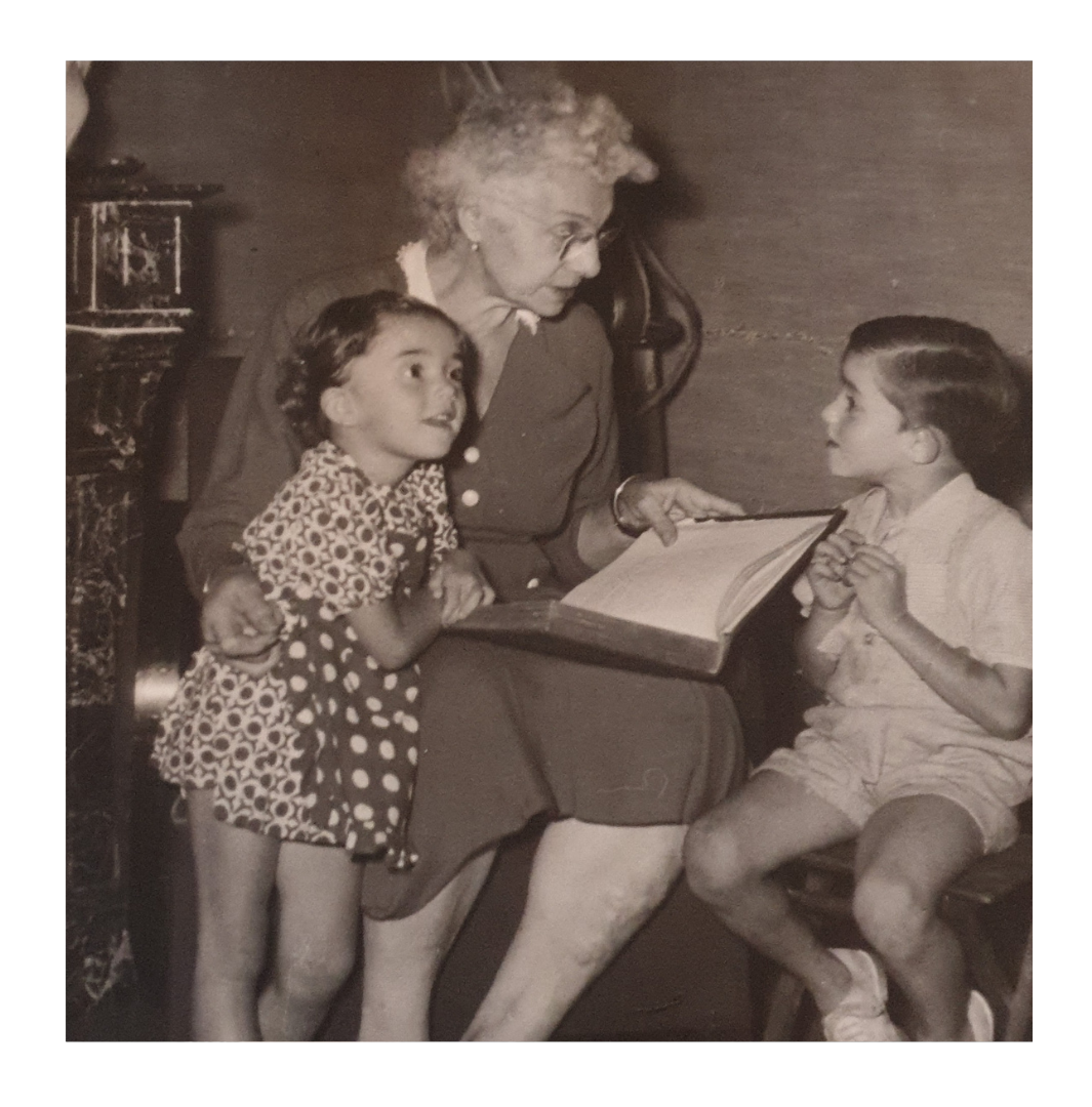 Une femme âgée lit un livre à deux enfants à l'intérieur, tous assis très près les uns des autres.