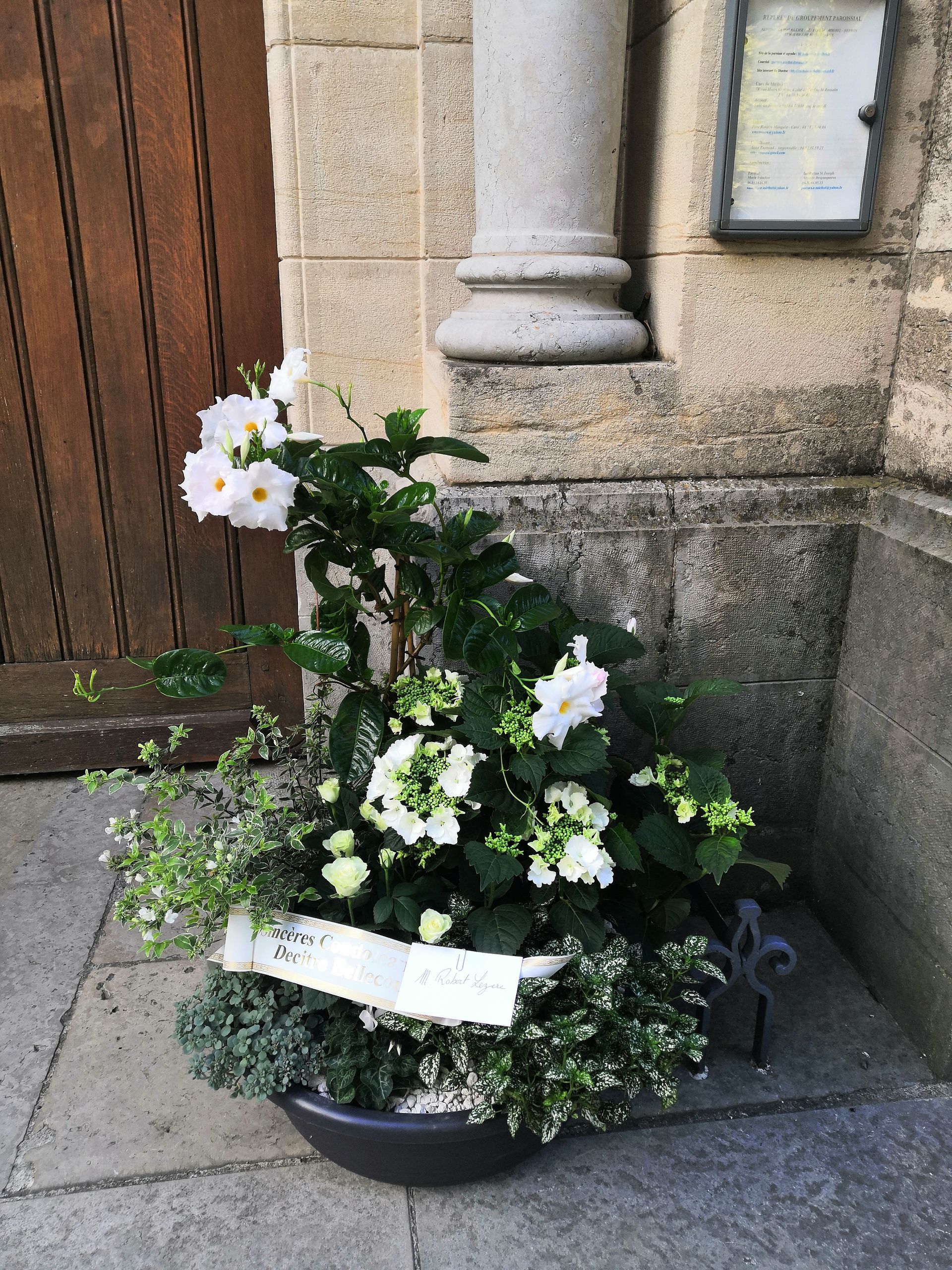 Des fleurs blanches en pot, ornées d'un ruban, sont placées à côté de l'entrée d'un bâtiment en pierre.