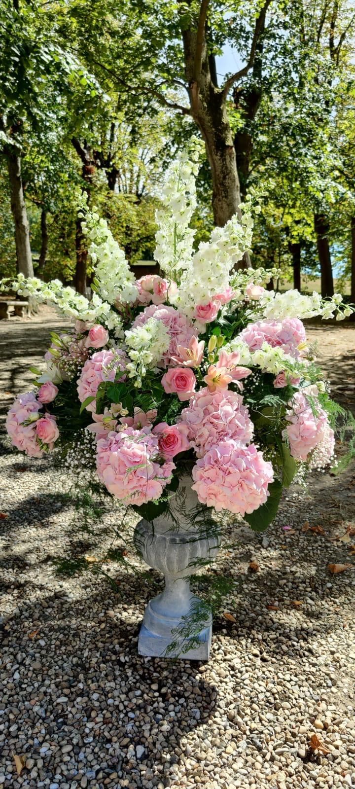Grande composition florale rose pastel et blanche dans un vase gris à l'extérieur, parmi les arbres et le gravier.