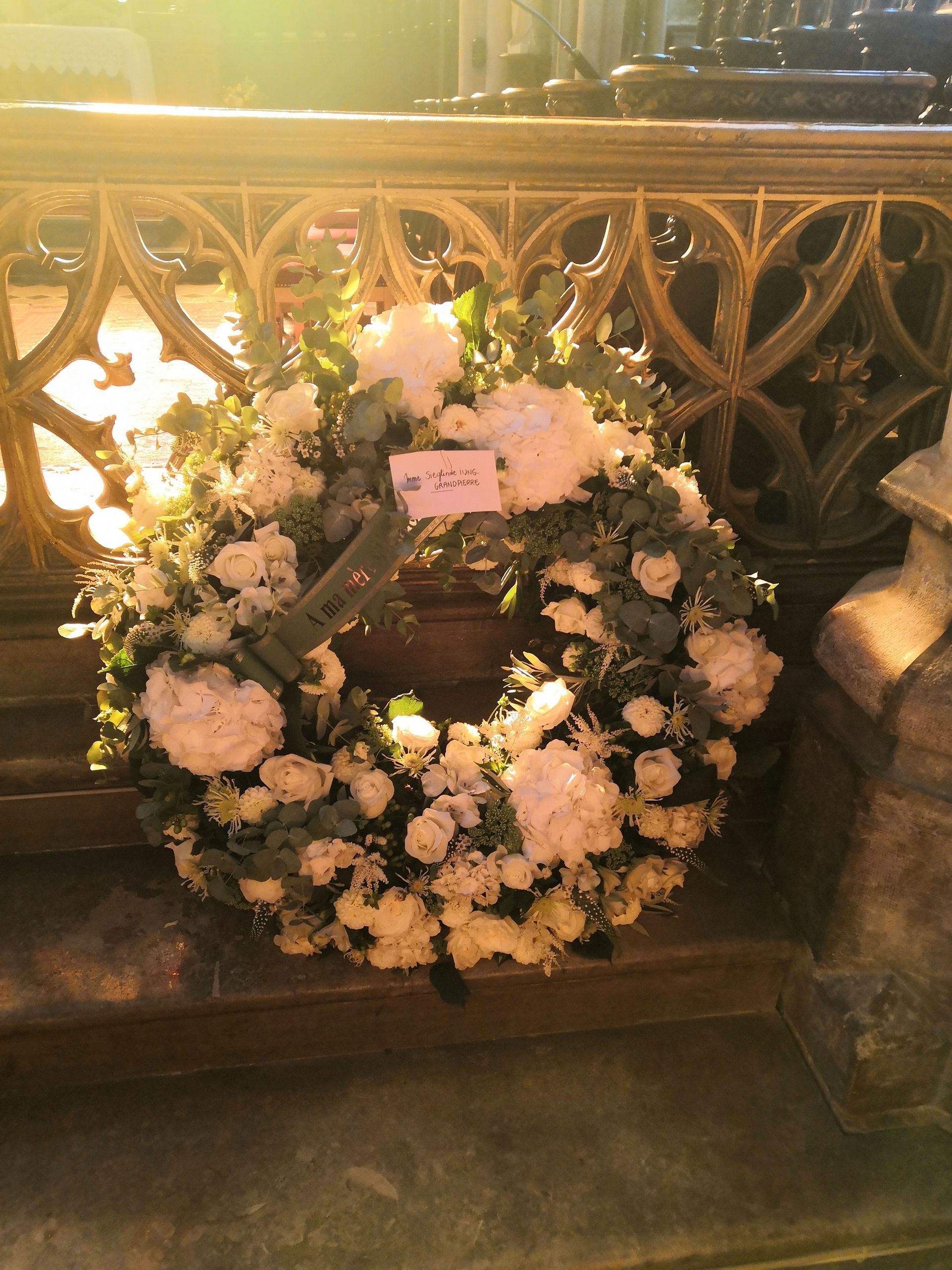 Couronne funéraire de fleurs blanches avec une carte rose, disposée devant une balustrade et un banc ornés.
