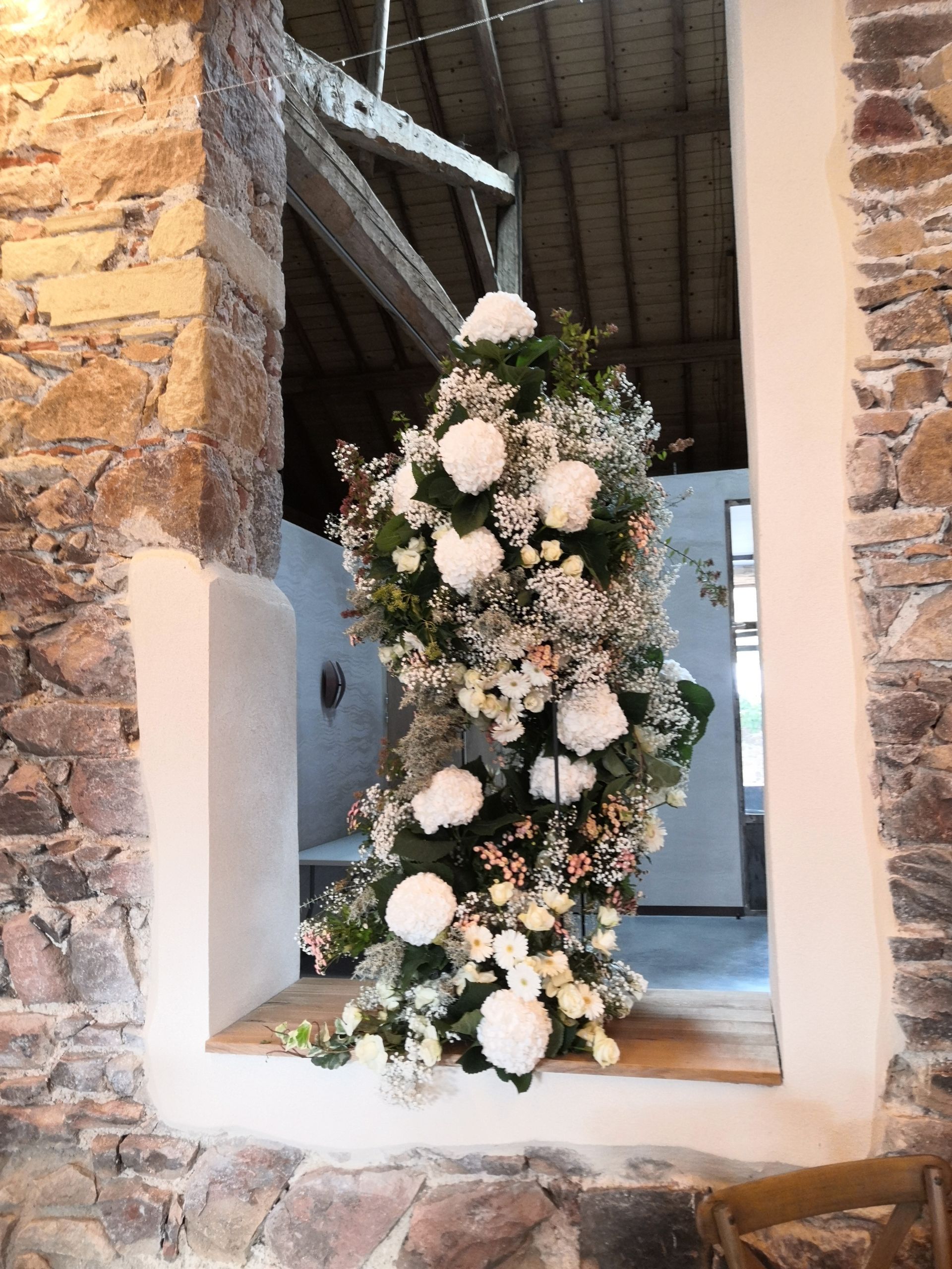 Grande composition florale blanche et rose poudré dans une ouverture de fenêtre en pierre d'un lieu événementiel intérieur