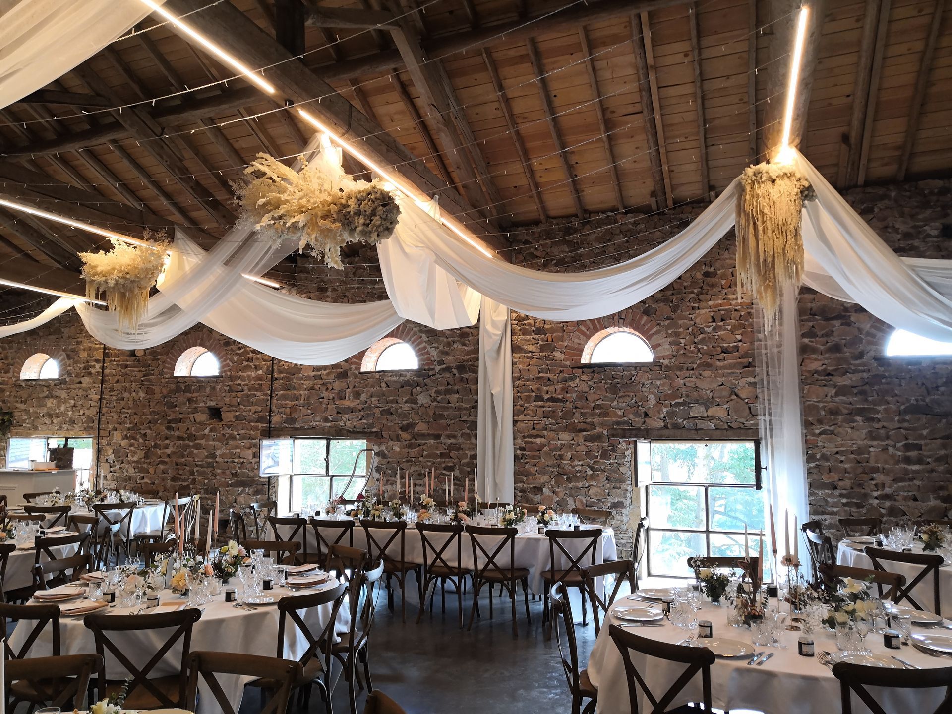 Salle de banquet rustique avec murs de briques, draperies blanches, tables en bois et lumières suspendues chaleureuses