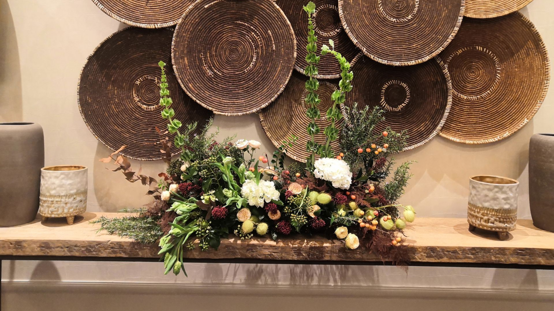 Composition florale décorative sur une table sous des paniers muraux tressés, avec des bougies de chaque côté.