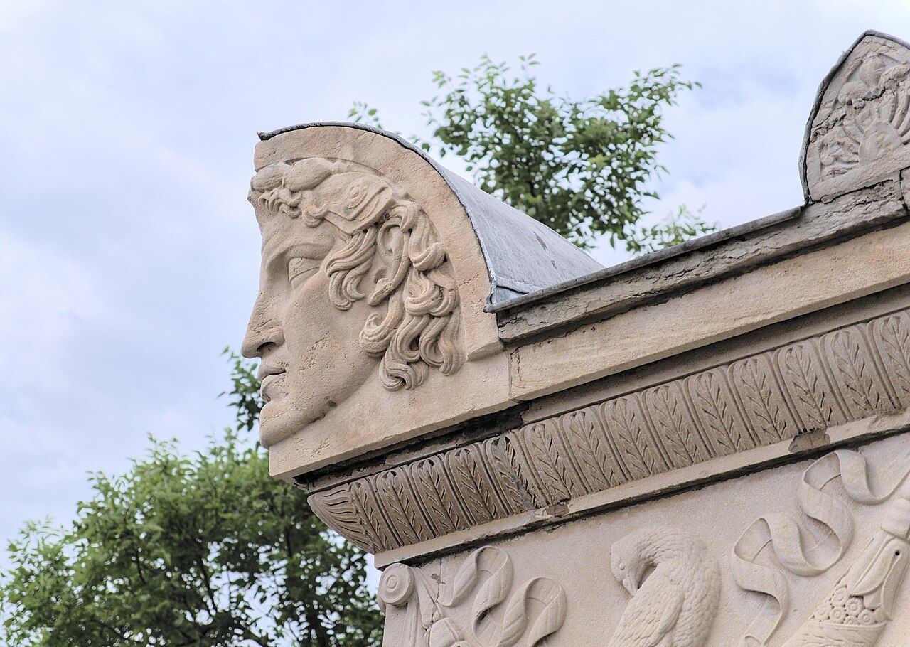 Bas-relief en pierre représentant un visage classique sur la corniche d'un bâtiment, se détachant sur un ciel nuageux et des arbres.