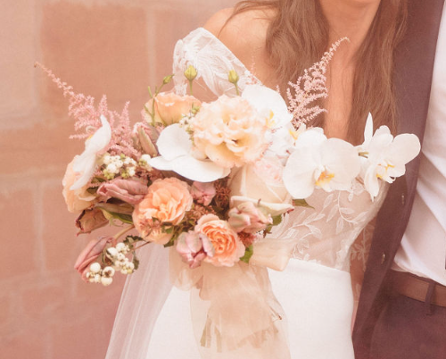 La mariée tient un bouquet pastel de fleurs blanches, pêche et roses dans un décor rose tendre