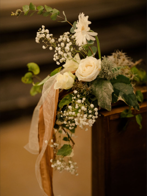 Composition florale crème et blanche avec verdure et rubans sur un piédestal en bois, sur fond sombre.