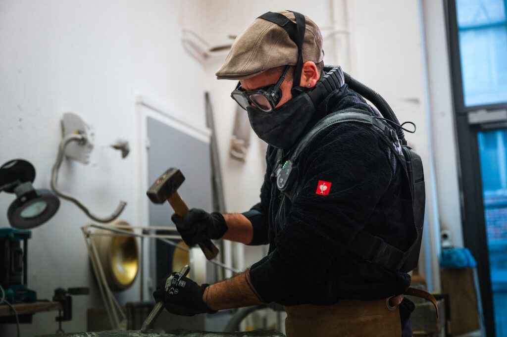 Ein Arbeiter, der in einer Werkstatt mit Hammer und Werkzeug arbeitet, trägt eine Kappe, eine Schutzbrille und eine Gesichtsmaske.