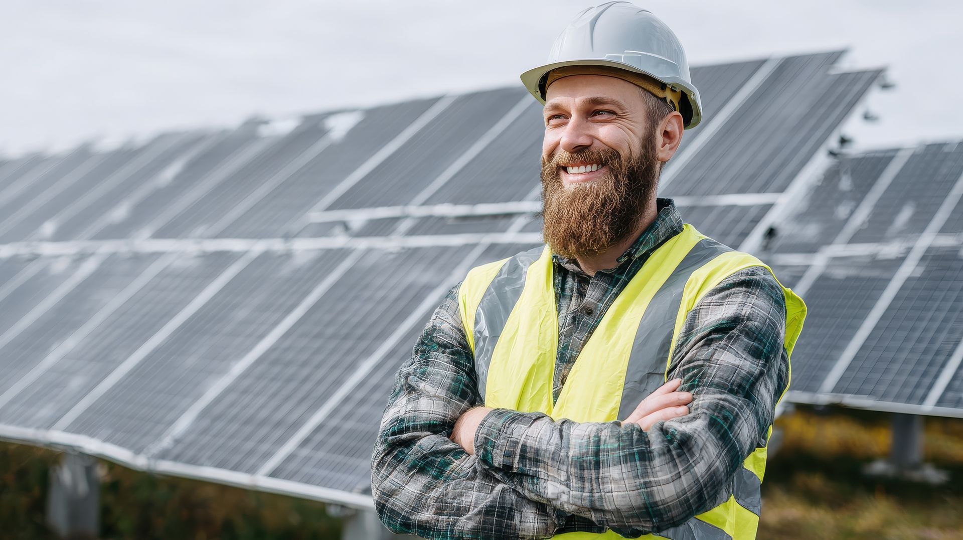 Un homme barbu, portant un gilet de sécurité et un casque de chantier, sourit, les bras croisés, devant des panneaux solaires.
