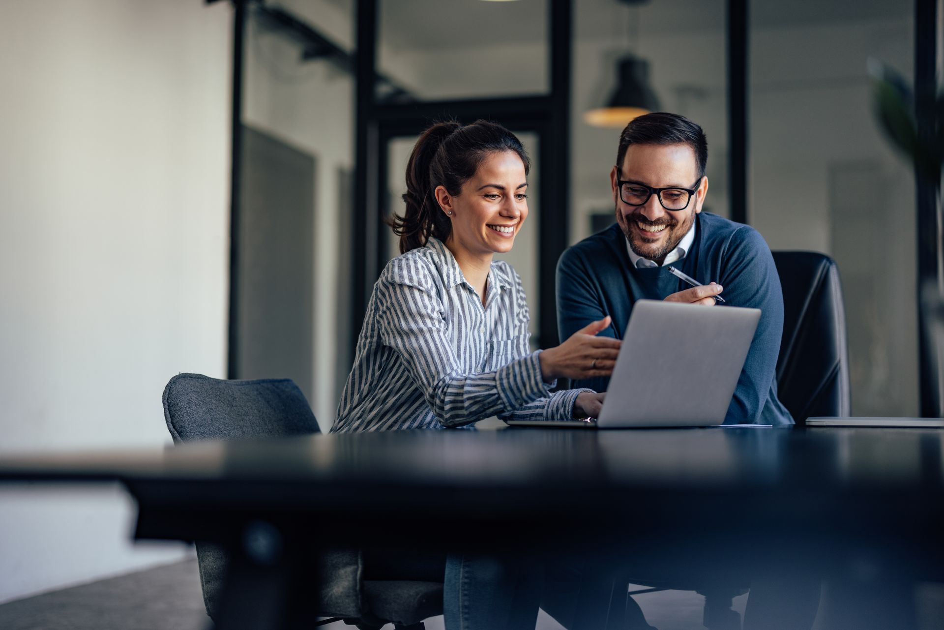 Deux professionnels souriants, les yeux rivés sur leur ordinateur portable, dans un environnement de bureau moderne.