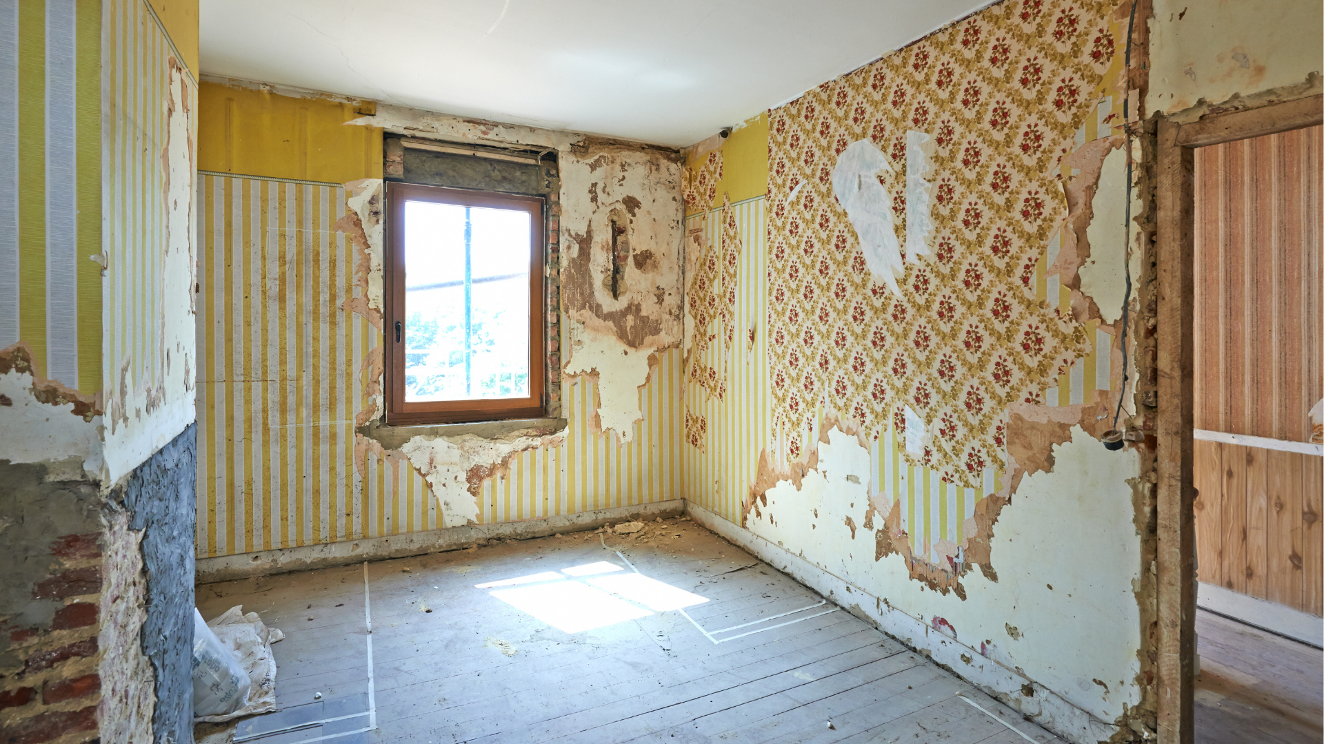 Pièce avec papier peint décollé, fenêtre en bois et murs apparents, probablement en cours de rénovation.