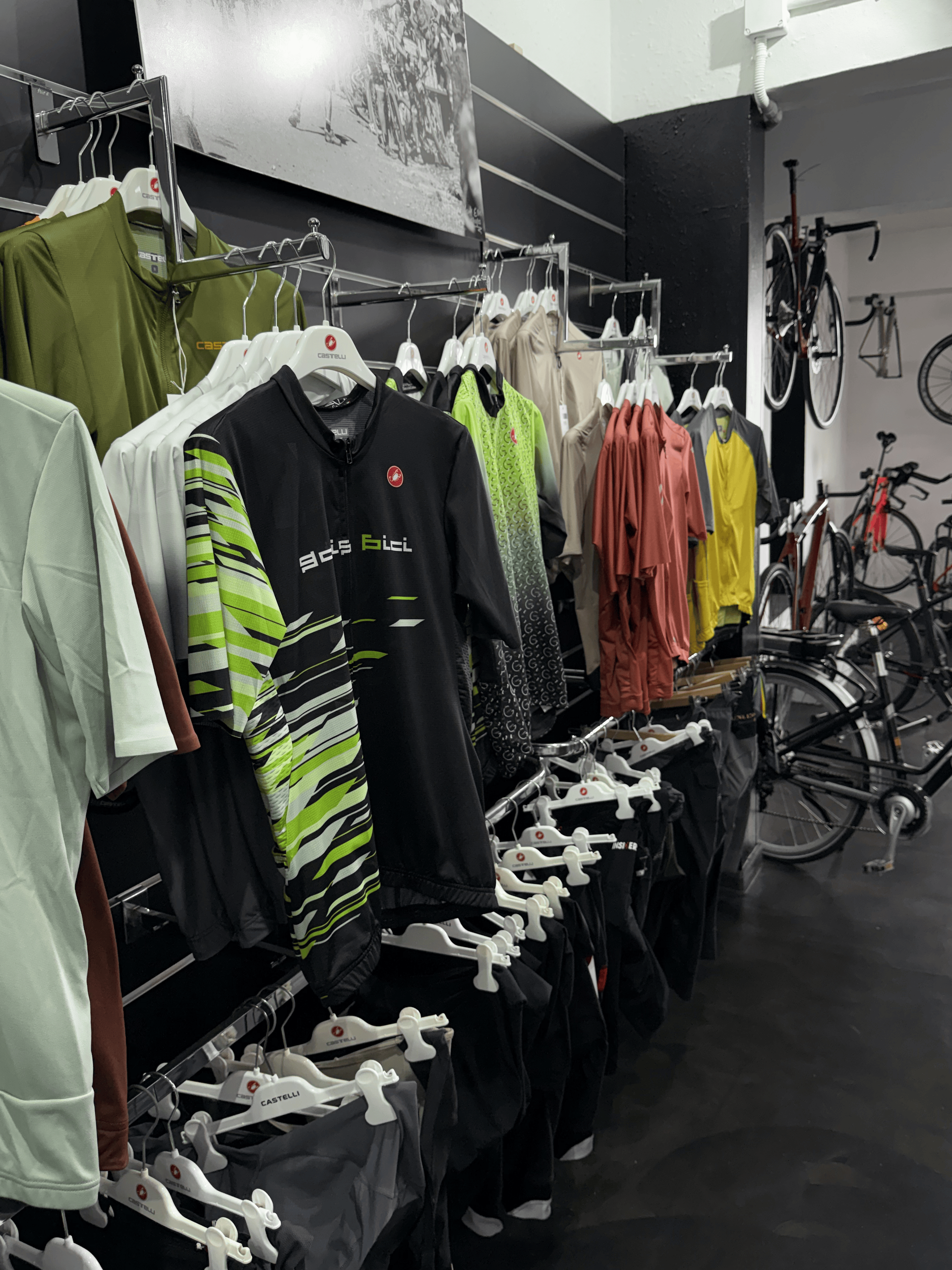 Tienda de ropa con percheros llenos de camisetas coloridas y equipamiento para ciclistas junto a bicicletas en una sala de exposición con suelo negro.