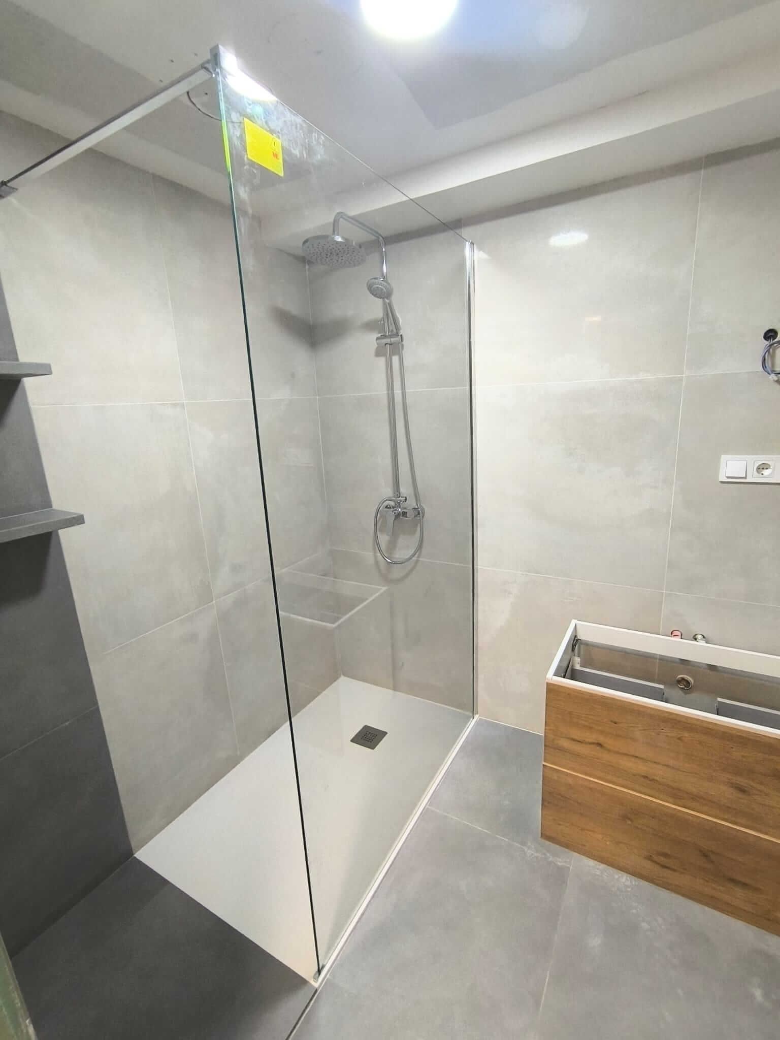 Baño moderno con paredes de azulejos grises, mampara de ducha de cristal y tocador de madera.