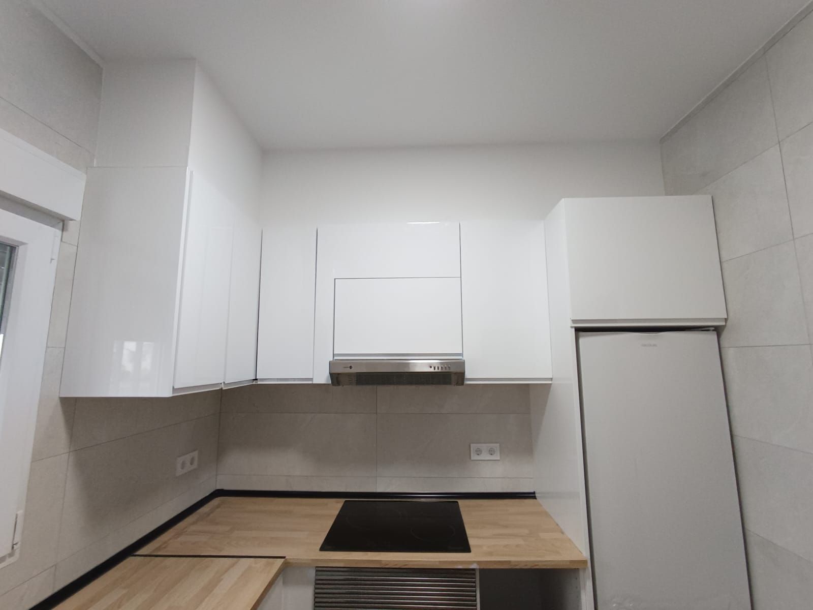 Cocina blanca moderna con encimeras de madera clara, campana extractora de acero inoxidable y refrigerador.