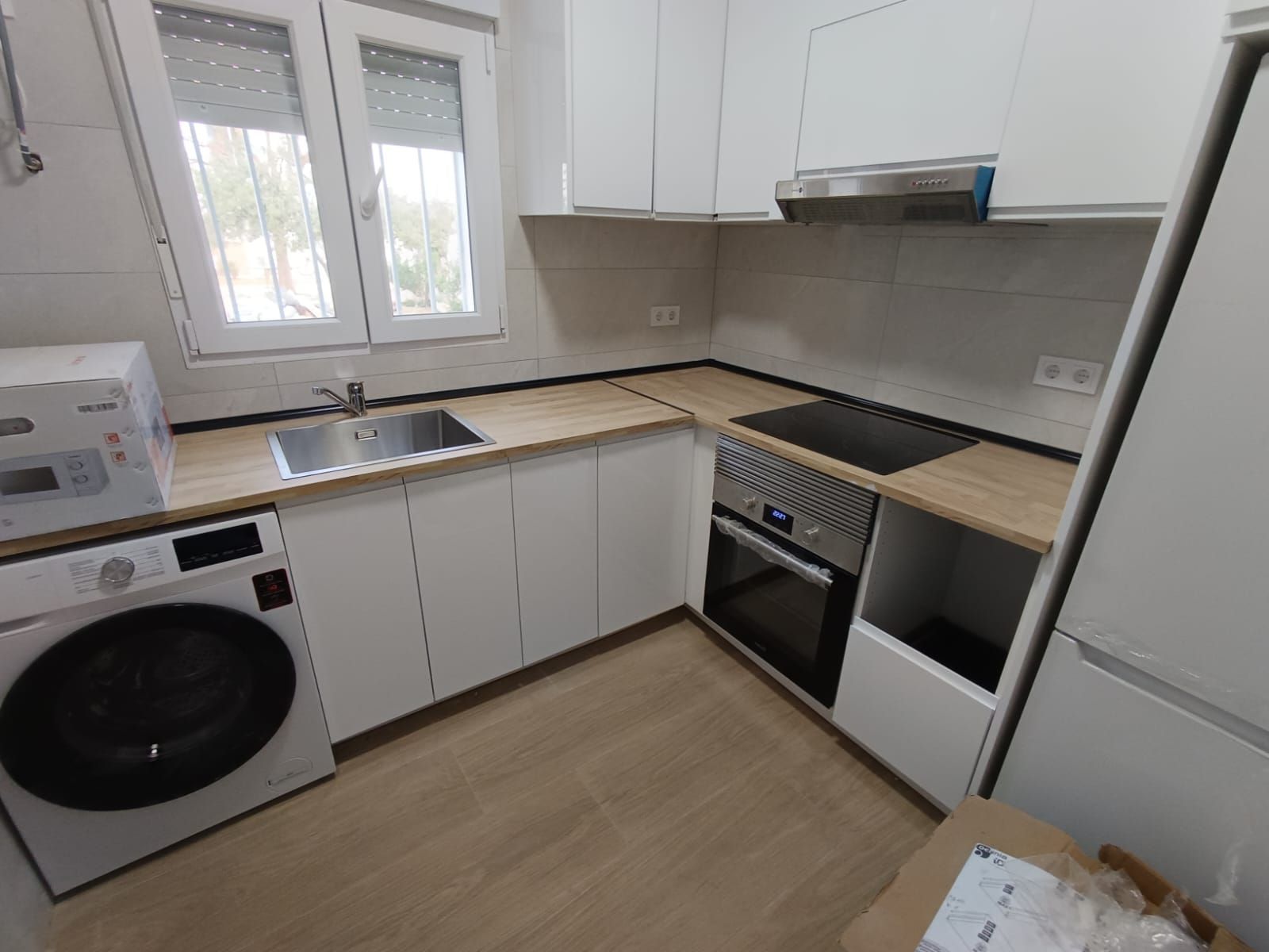 Cocina pequeña y blanca con encimeras de madera, electrodomésticos de acero inoxidable y una ventana.