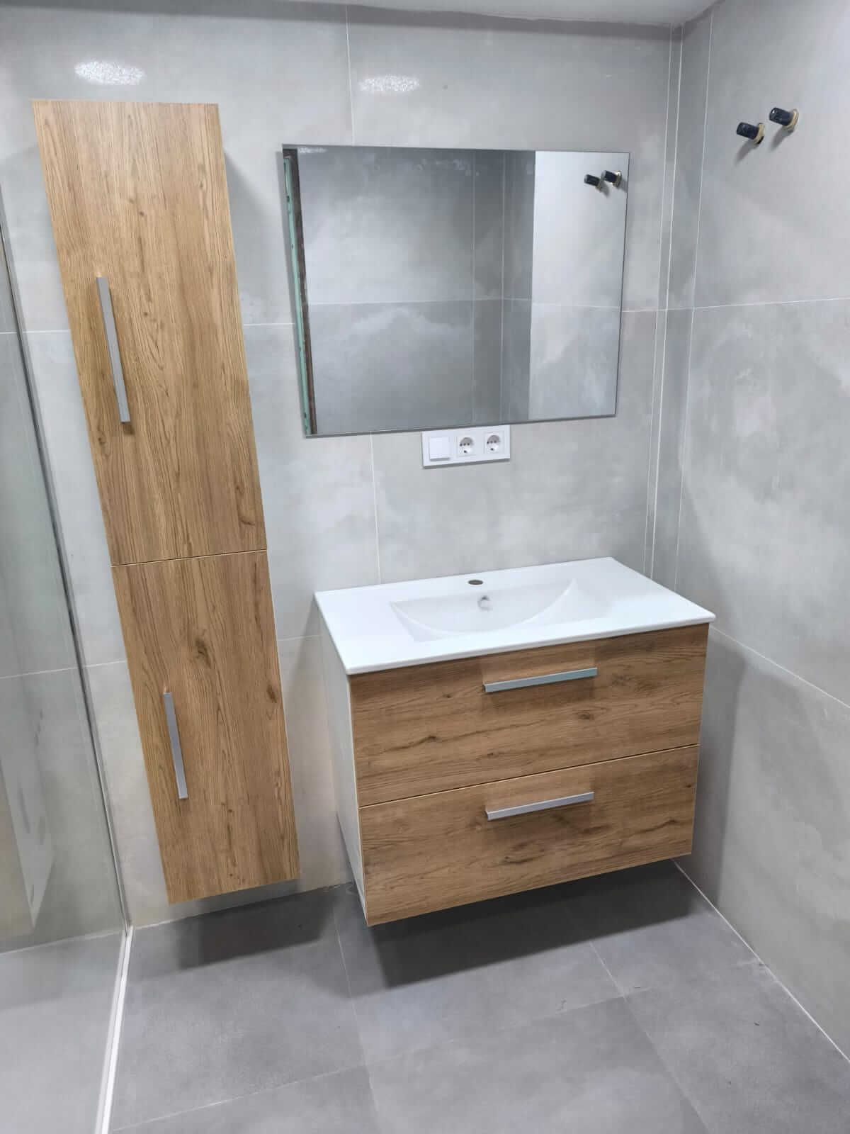 Baño moderno con tocador de madera y armario alto. Espejo cuadrado y paredes de azulejos grises.