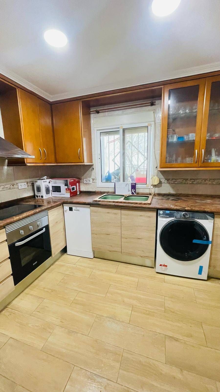 Cocina con gabinetes de madera, electrodomésticos y ventana. Pisos y encimeras beige.