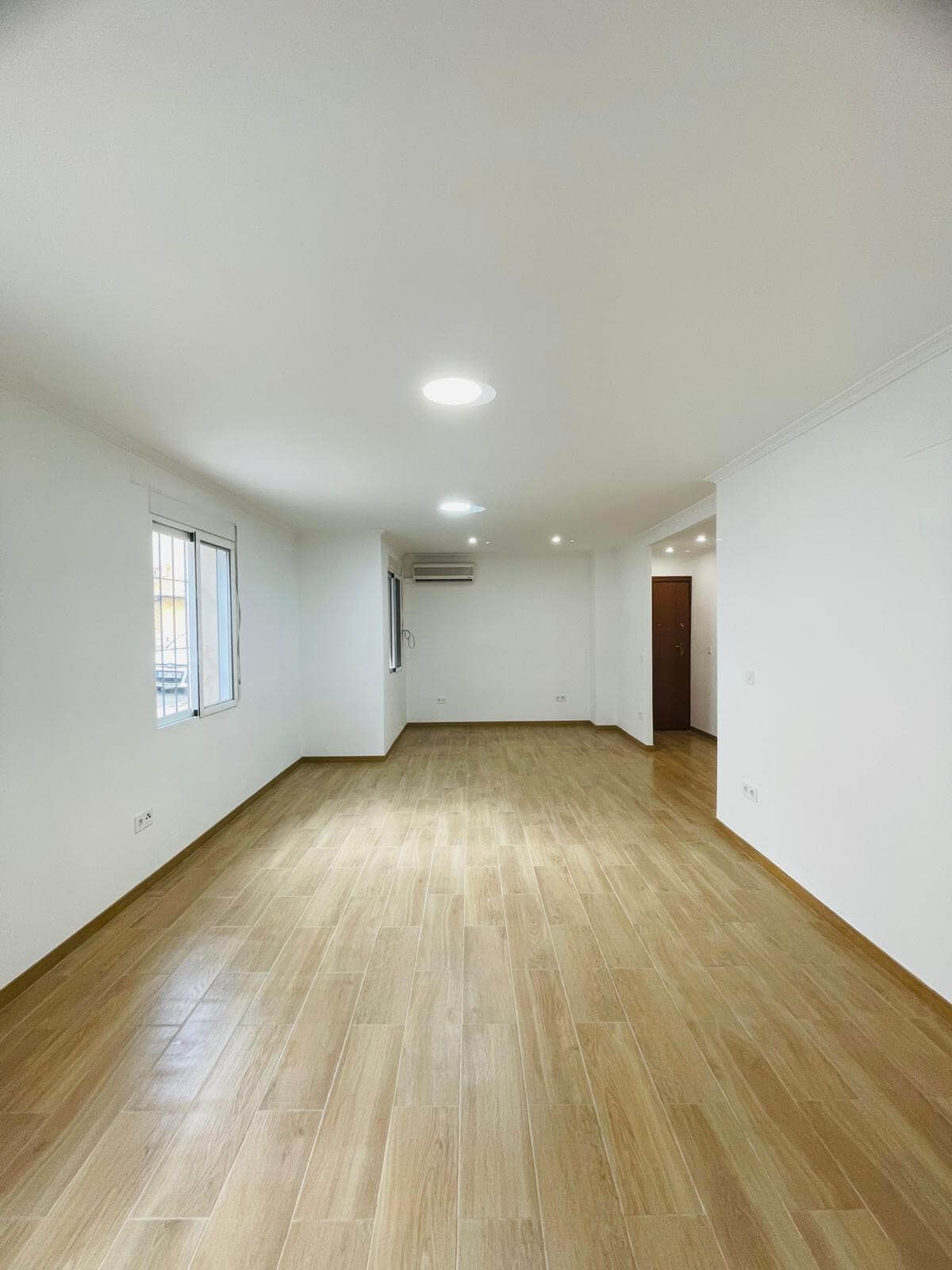 Habitación vacía y bien iluminada, con pisos de madera clara, paredes blancas y luces empotradas en el techo.