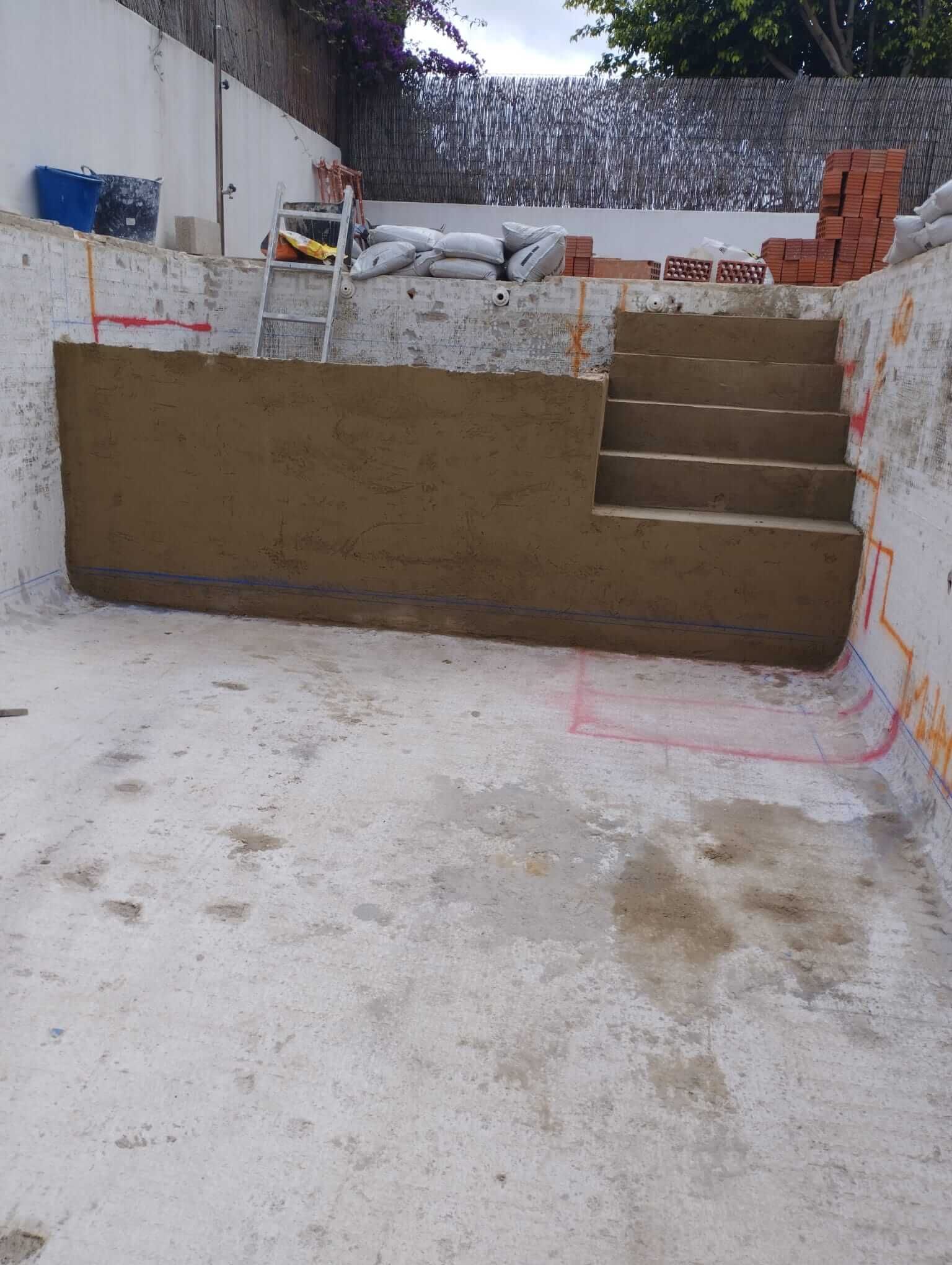 Construcción de piscina: piscina de hormigón con escalones marrones, pared marrón, materiales de construcción al fondo.