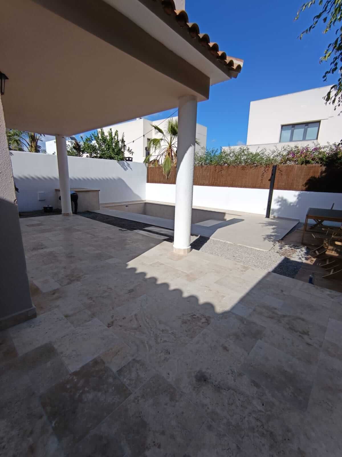 Un patio cubierto con suelo de piedra, una columna blanca y vistas a una valla y a las casas vecinas.