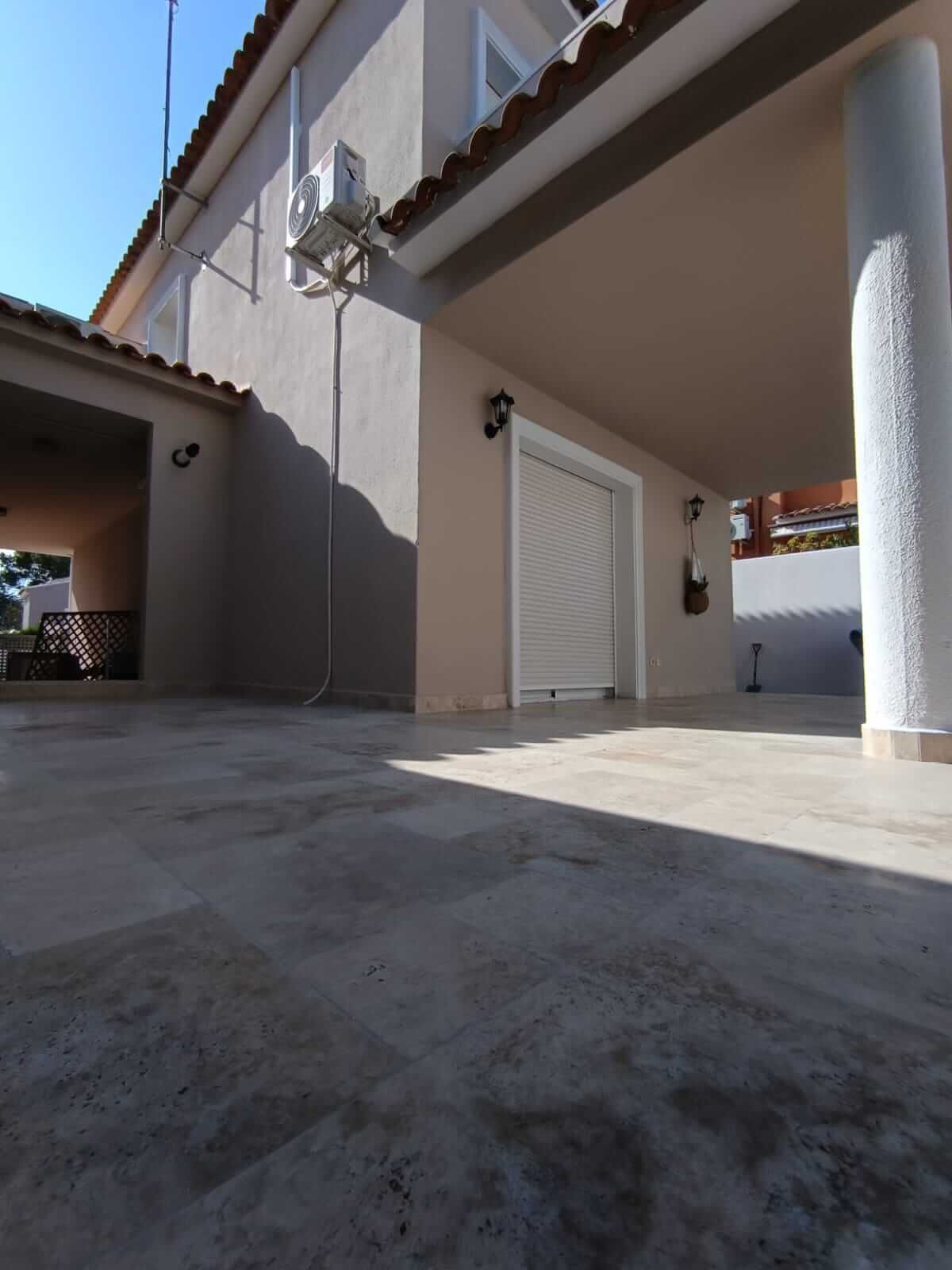 Casa de dos plantas de color beige con porche cubierto, contraventanas blancas y un gran patio de piedra de color claro.