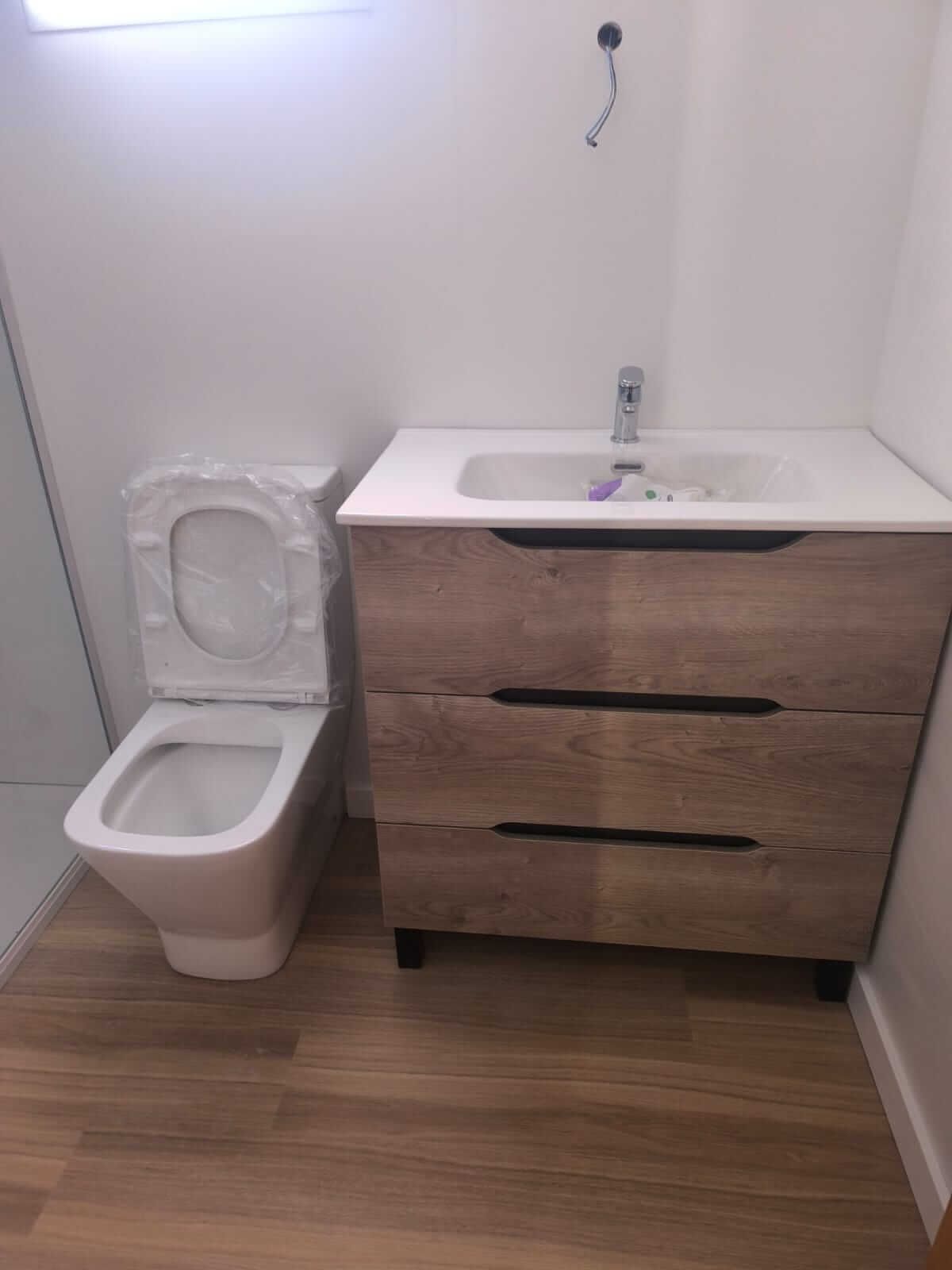 Baño con inodoro, lavabo y mueble de madera; suelo marrón claro y paredes blancas.