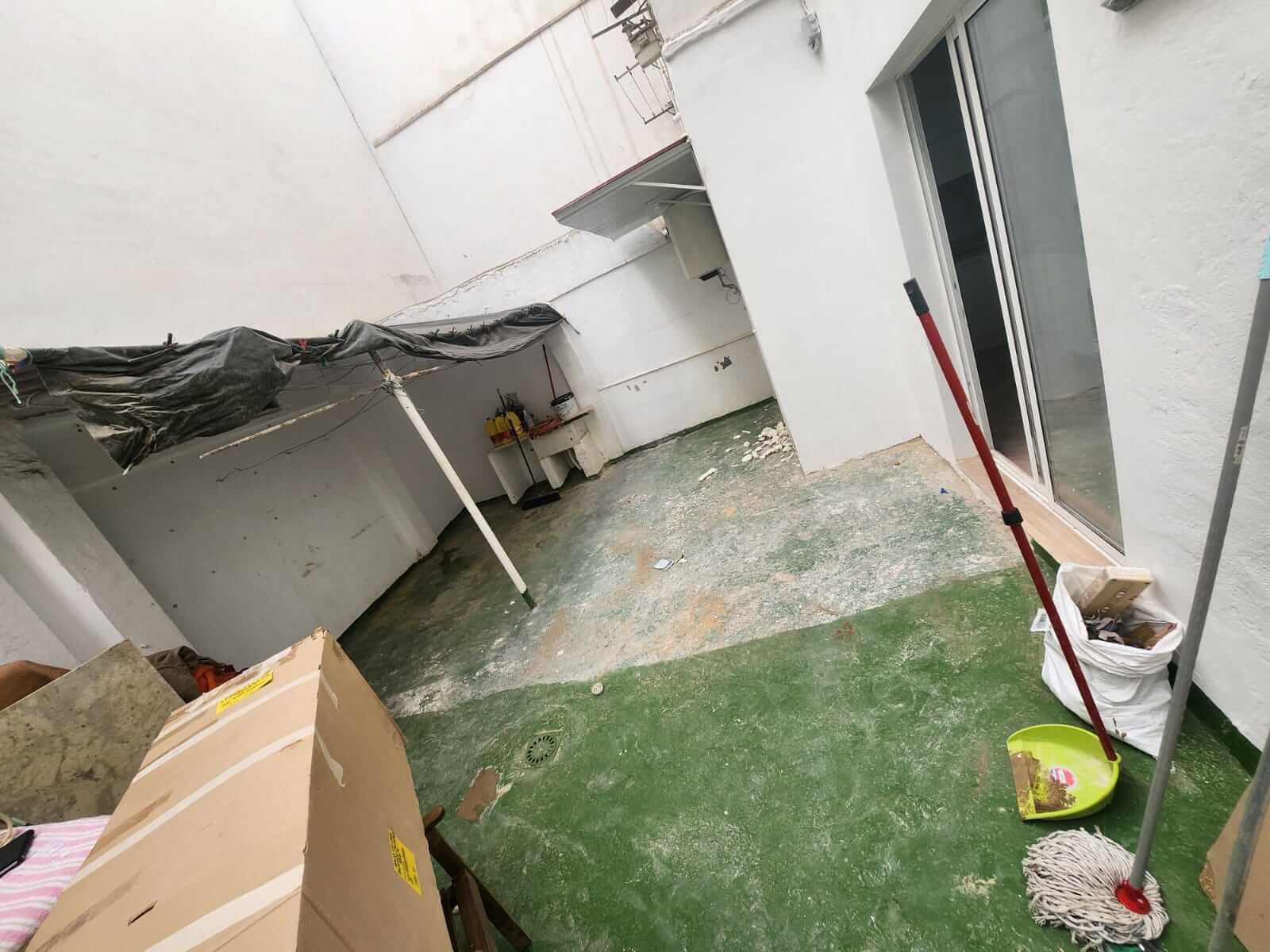 Habitación desordenada y abarrotada con piso verde, puerta abierta, cajas de cartón y artículos de limpieza.