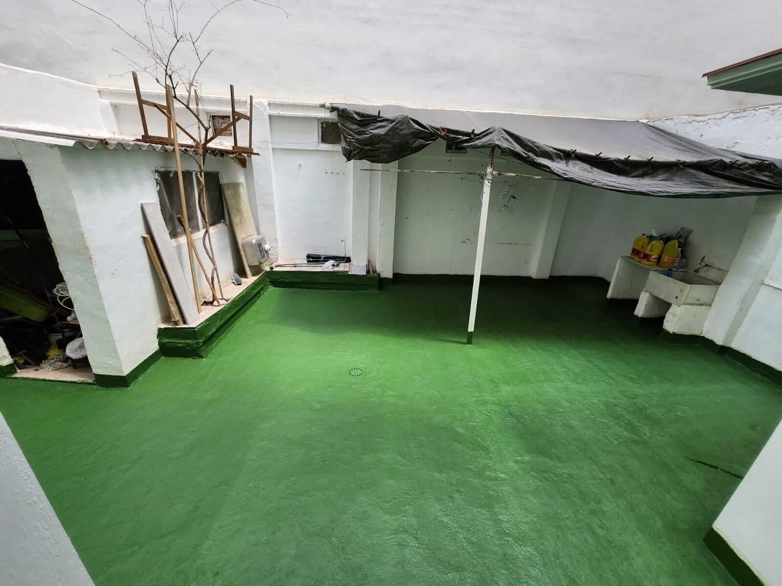 Patio interior con suelo verde y paredes blancas. Hay un tendedero bajo una lona negra; varios objetos y un pequeño árbol.