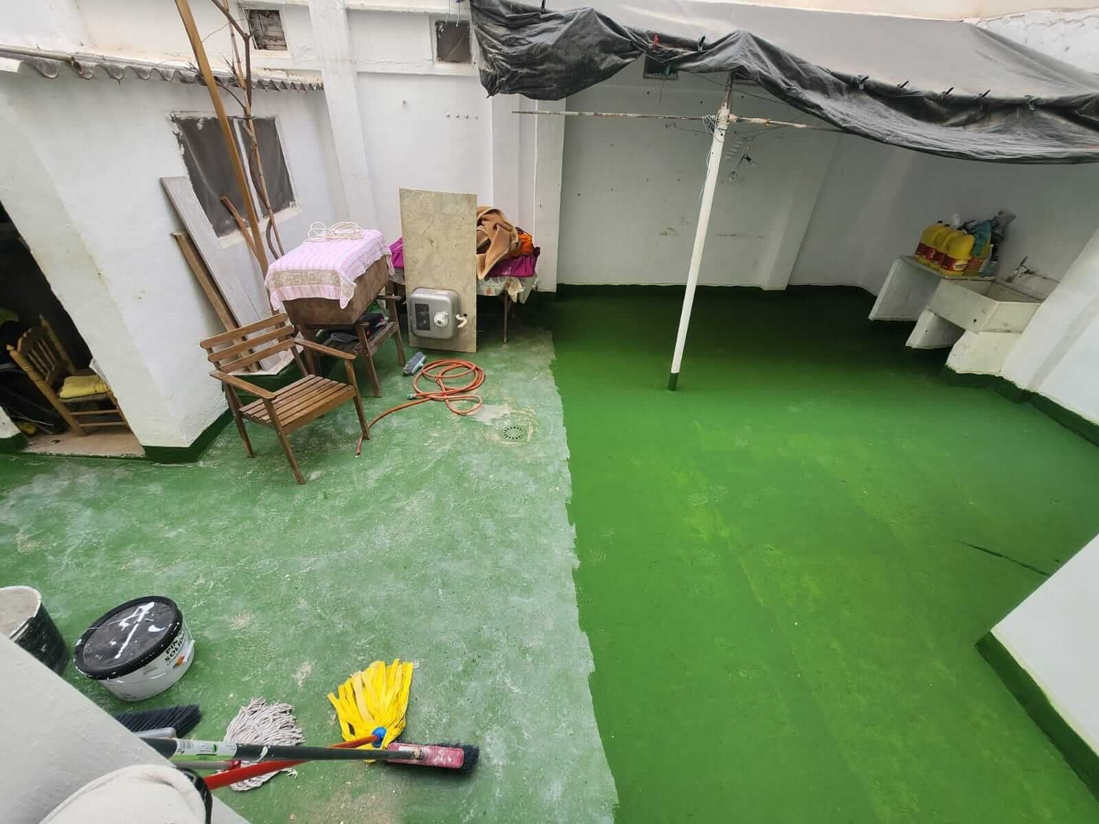 Un patio medio pintado de verde. Hay muebles, herramientas y una lona para dar sombra.