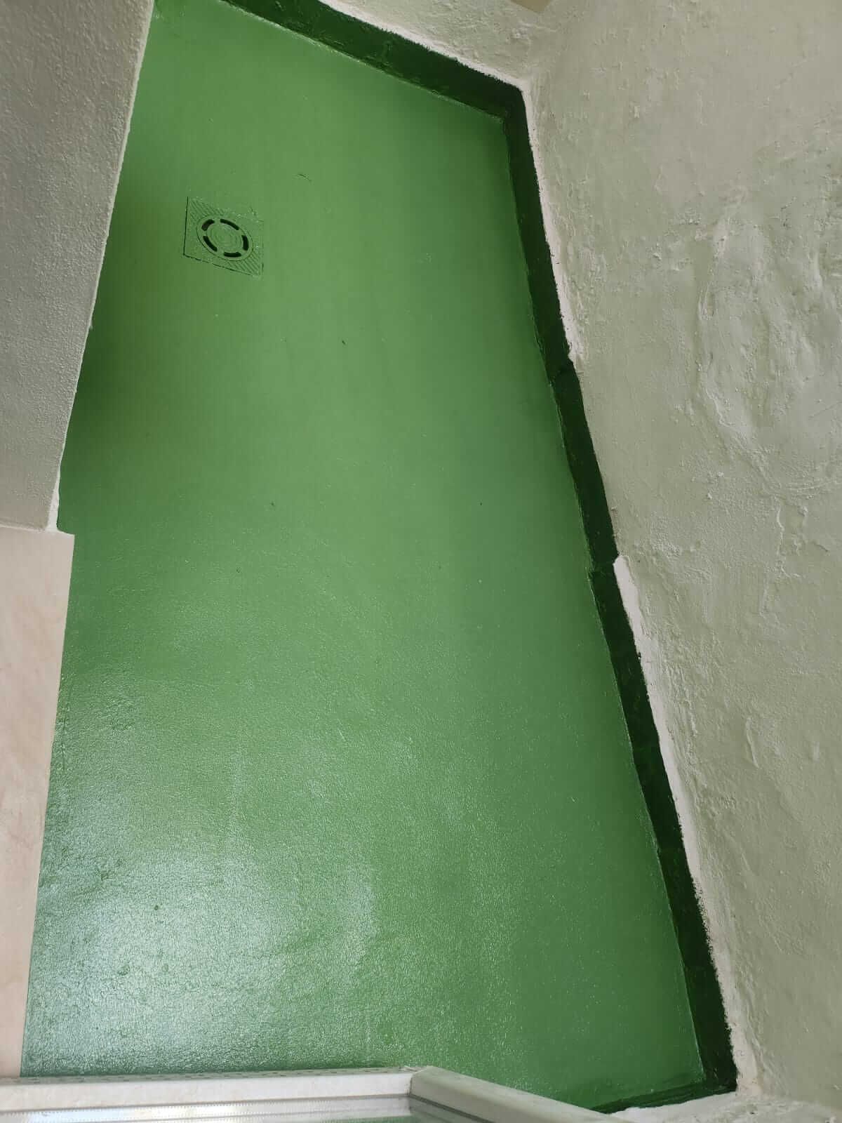 Suelo de ducha pintado de verde con bordes verde oscuro y desagüe. Paredes blancas rodean el suelo.