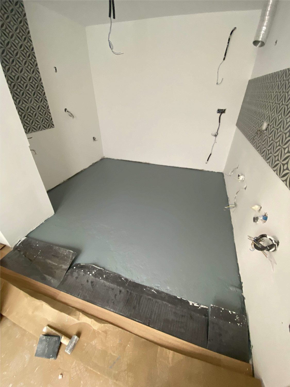 Baño en construcción con piso gris, paredes blancas, azulejos estampados y cableado expuesto.