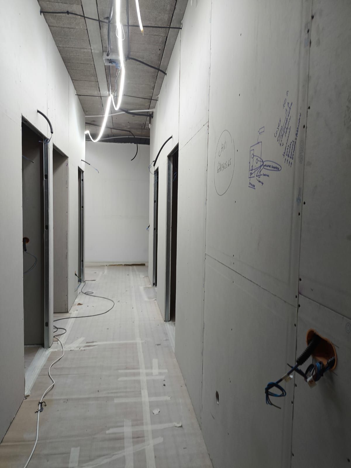 Un pasillo en construcción con paneles de yeso y cableado sin terminar, varias puertas en el lado izquierdo.