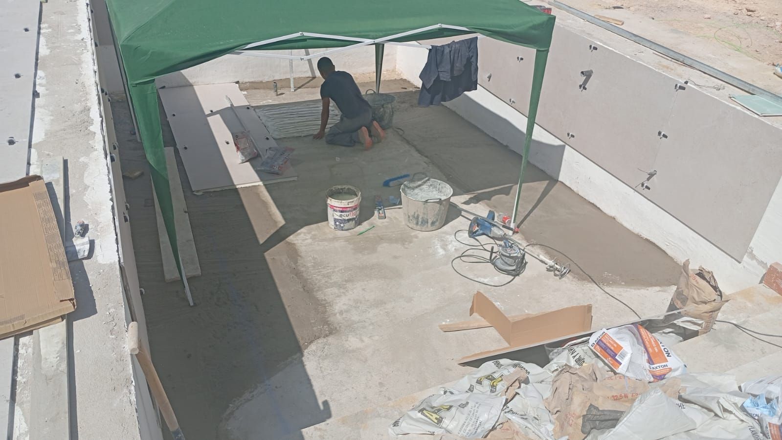 Hombre trabajando bajo un dosel verde sobre una superficie de hormigón; herramientas y materiales esparcidos alrededor.