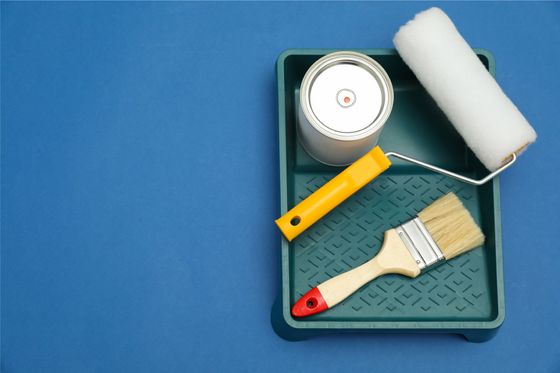 Suministros de pintura en una bandeja: lata de pintura, rodillo, pincel, todo sobre un fondo azul.