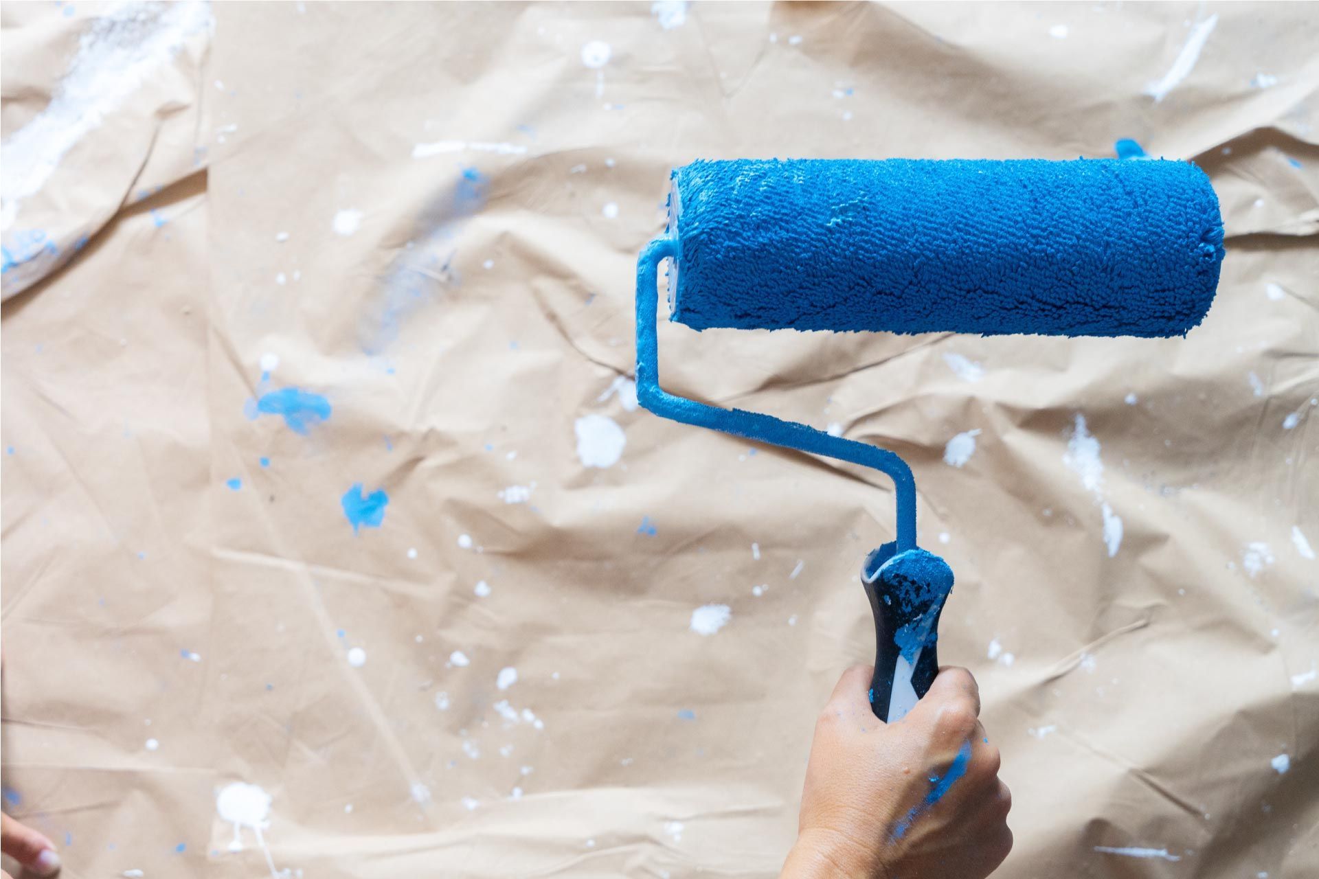 Mano sosteniendo un rodillo de pintura azul sobre una superficie de papel marrón, salpicada con pintura azul y blanca.