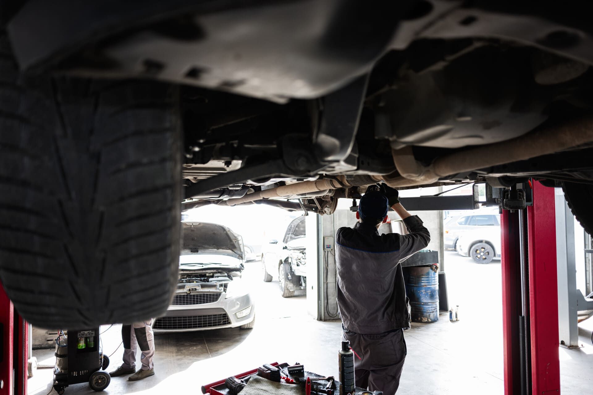 Mecánico trabajando debajo de un coche en un taller mecánico luminoso con vehículos al fondo.