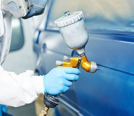 Un trabajador con guantes rocía la carrocería azul de un coche con una pistola de pintura en un taller mecánico.