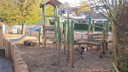 Spielplatzanlage aus Holz mit Rutsche und Klettergerüsten, eingebettet in eine mit Sand bedeckte, von Bäumen umgebene Fläche.
