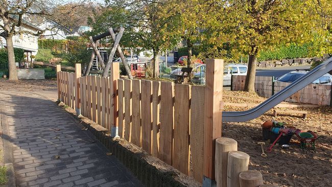 Ein hölzerner Lattenzaun grenzt an einen Spielplatzweg; im Hintergrund sind Bäume, Herbstlaub und eine Rutsche zu sehen.