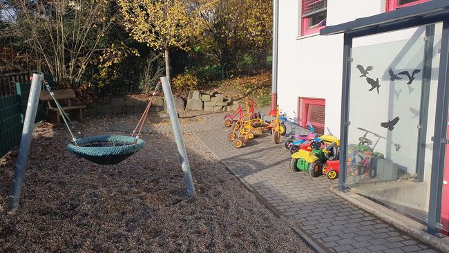 Ein Spielbereich im Freien mit einer kreisförmigen Hängeschaukel und verschiedenen bunten Spielzeugfahrzeugen in der Nähe.