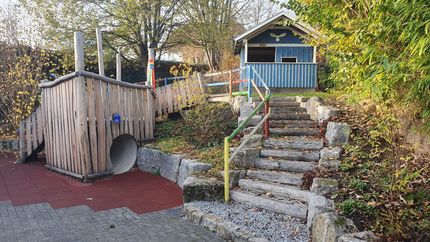 Ein hölzernes Klettergerüst, einem Kriechtunnel und einer Steintreppe, die zu einem blauen Schuppen führt.